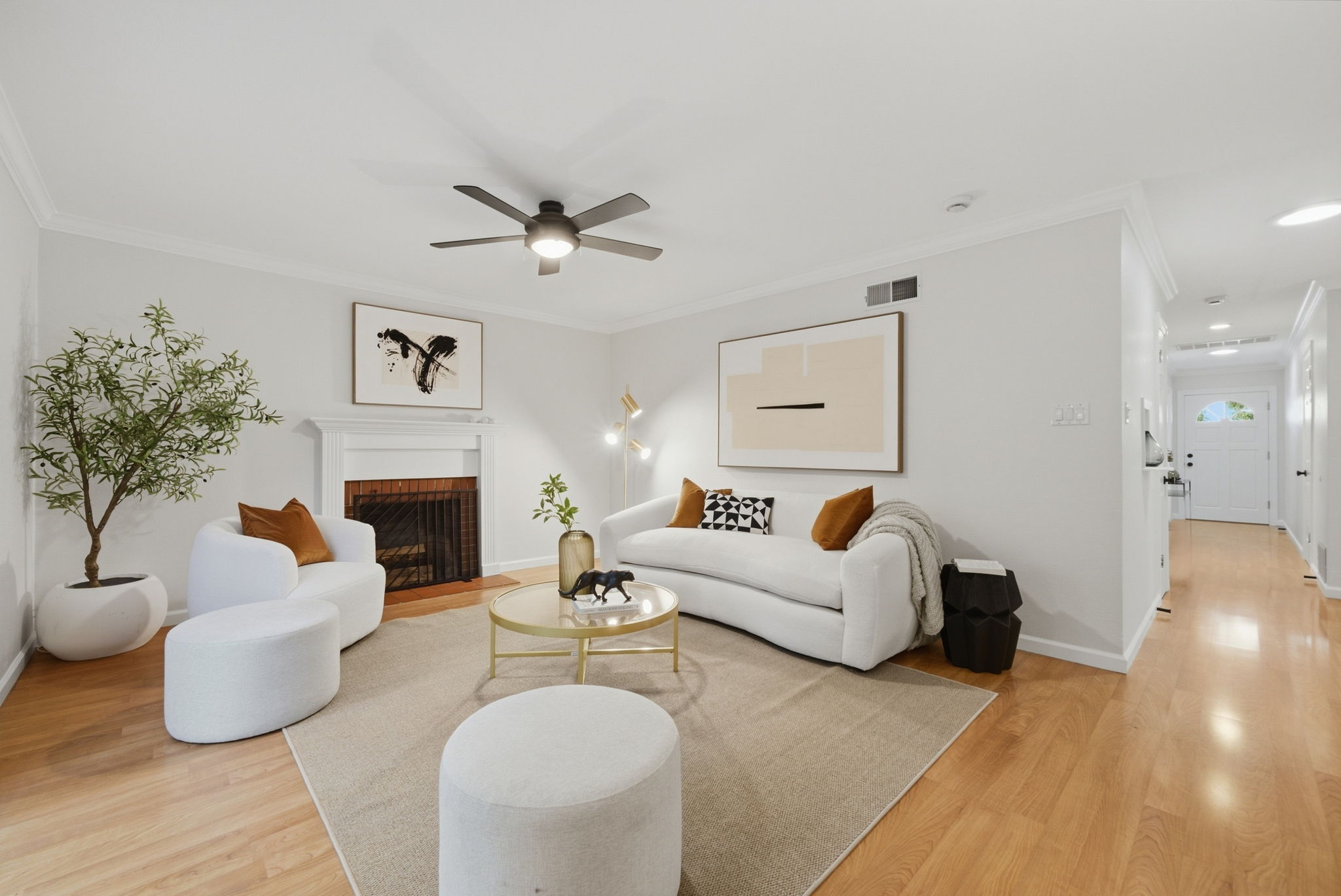 Modern living room with light wood flooring, white walls, a white sofa with brown and black patterned cushions, a matching white armchair with a brown cushion, and two round white ottomans on a large beige rug. A round gold coffee table holds a vase with greenery and a decorative panther figure. There is a black ceiling fan with five blades and a light in the center. A white fireplace with a dark interior is on the left wall under abstract black and white artwork. A tall green potted plant stands beside the armchair. On the right wall above the sofa is a large framed abstract beige and black art piece, next to a modern gold floor lamp. A black side table with a book is beside the sofa; a hallway with light wood flooring and white doors extends to the front door.