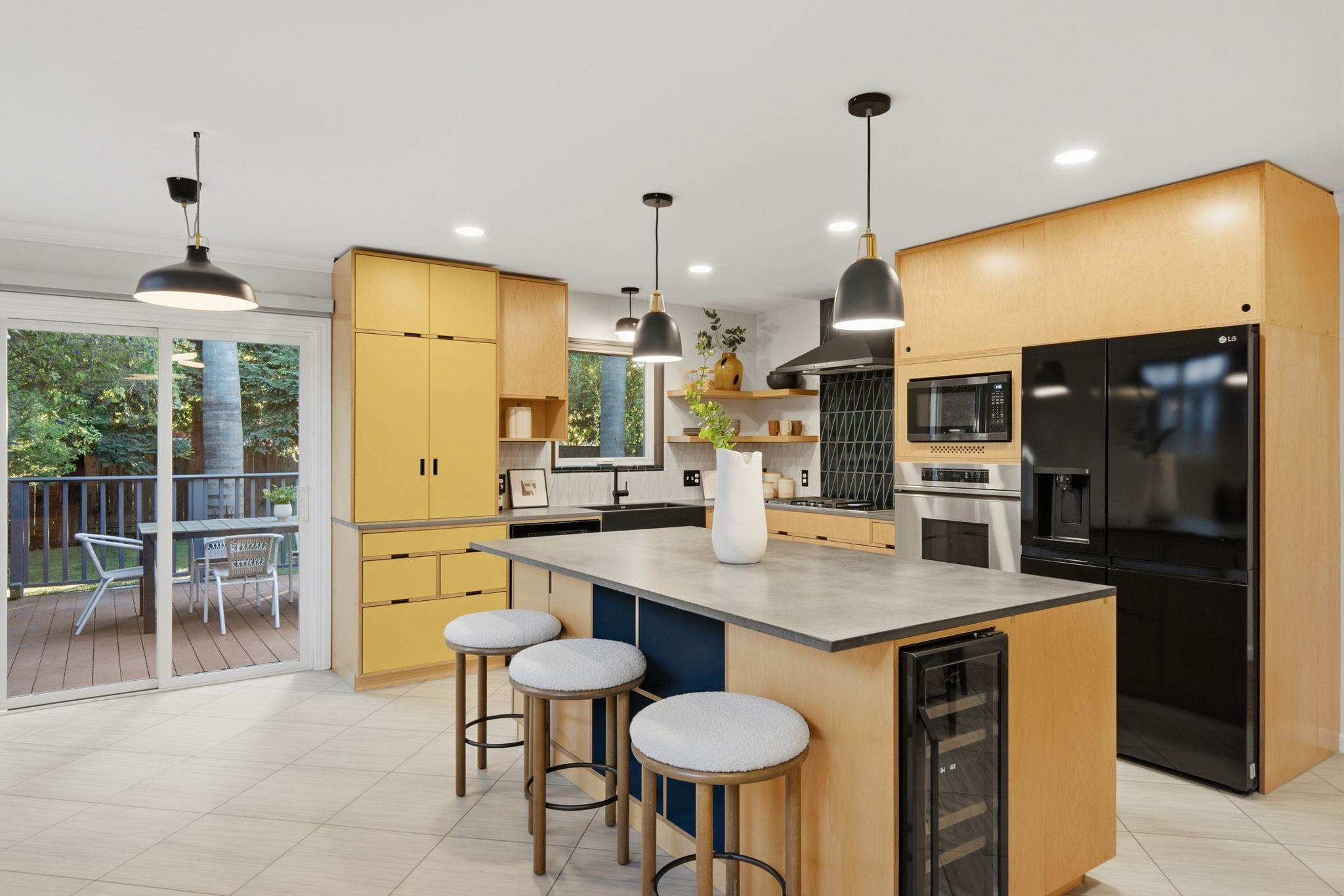 Modern kitchen with a large island featuring a gray countertop and three round stools with white cushions, yellow and natural wood cabinetry, black appliances including a refrigerator, oven, and microwave, pendant lights above the island, and a sliding glass door leading to an outdoor patio with seating and green trees visible outside.
