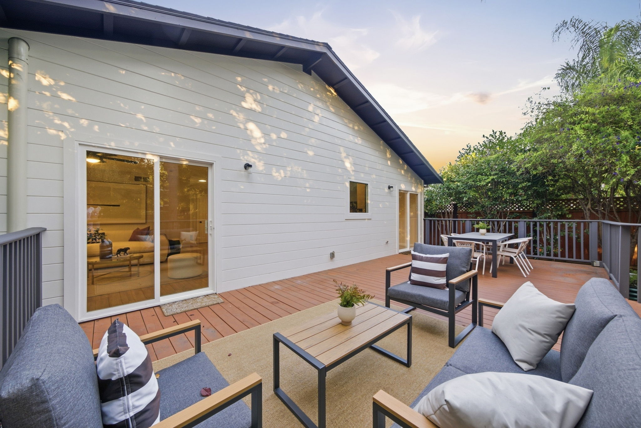 Outdoor wooden deck attached to a house with grey patio furniture including sofas and chairs with cushions, a wooden coffee table with a plant, and a separate dining table with chairs, surrounded by railing and trees at sunset.