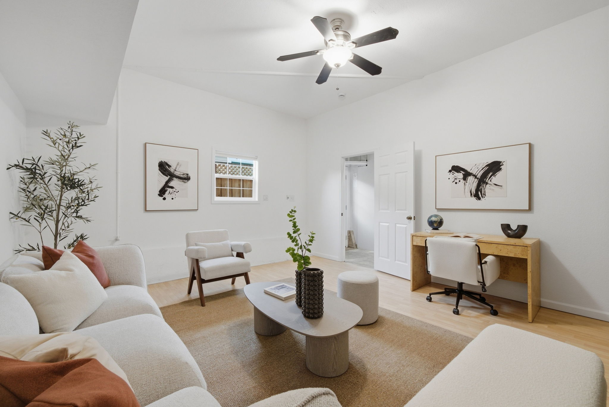 Bright modern living room with white walls and light wood floor featuring a white sofa with beige and rust pillows, a white armchair, beige ottoman, oval gray coffee table with a textured dark vase and a book, a tall plant in the corner, two abstract black and white paintings on the walls, a wooden desk with a rolling white chair, a globe, and decorative items, and a ceiling fan with five black blades turned on.