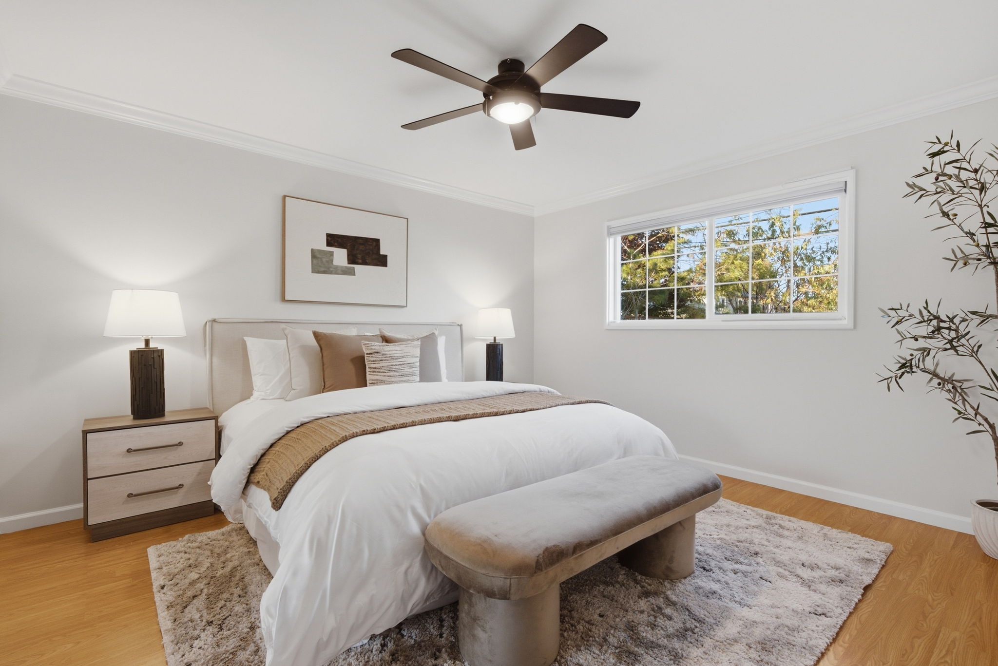 Bright bedroom with a queen bed dressed in white and beige bedding, a beige upholstered bench at the foot, two matching nightstands with lit lamps, abstract wall art above the bed, a ceiling fan with light, a large window showing green trees outside, a shaggy area rug, and a potted plant in the corner.