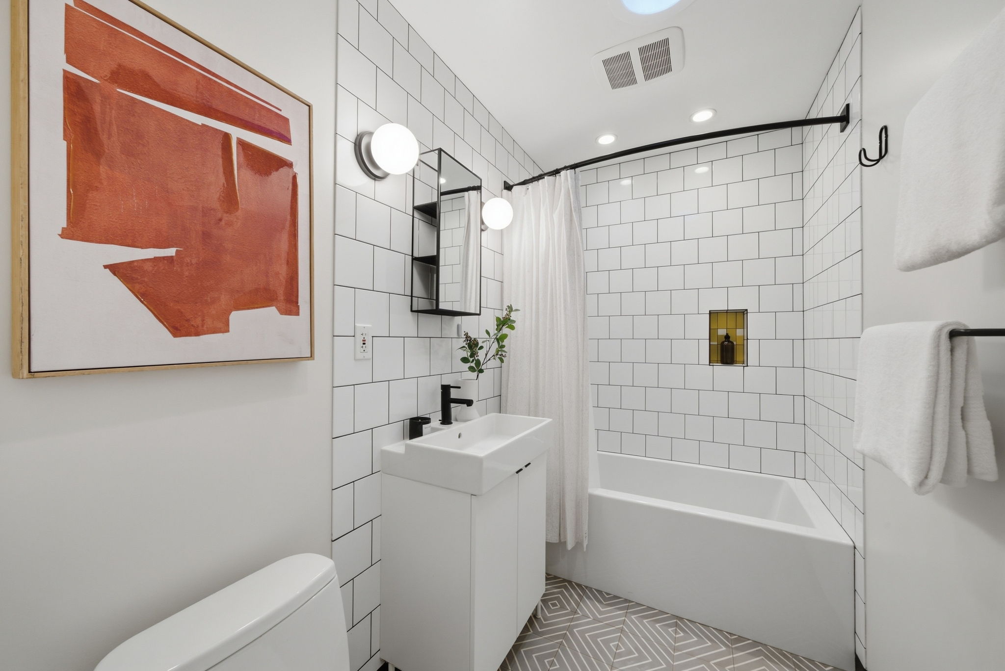Modern bathroom with white tiled walls and geometric patterned floor tiles, featuring a white bathtub with a shower curtain, a white vanity with a rectangular sink and black faucet, two round wall lights, a small mirror cabinet, white towels on a black towel rack, and a large abstract orange painting on the wall.