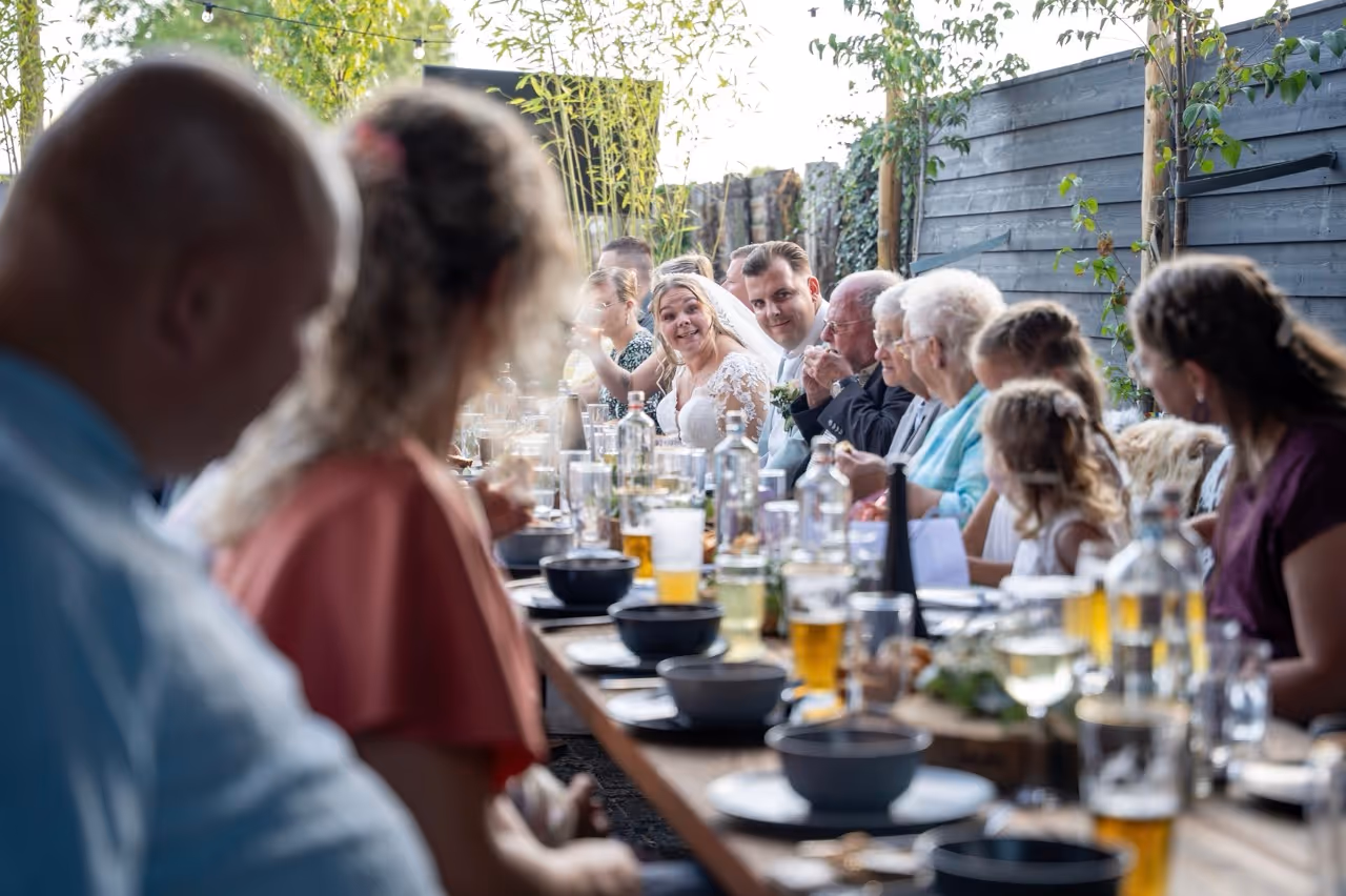 Bride and groom smiling at an outdoor wedding dinner with family and friends seated along a long table.
