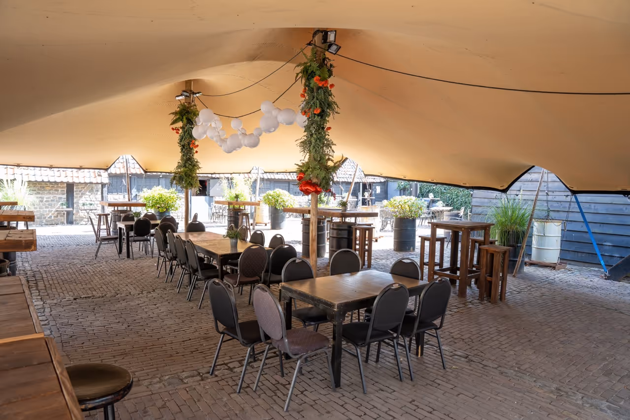 Outdoor seating area under a large beige canopy with tables, chairs, hanging flower arrangements, and cobblestone floor.