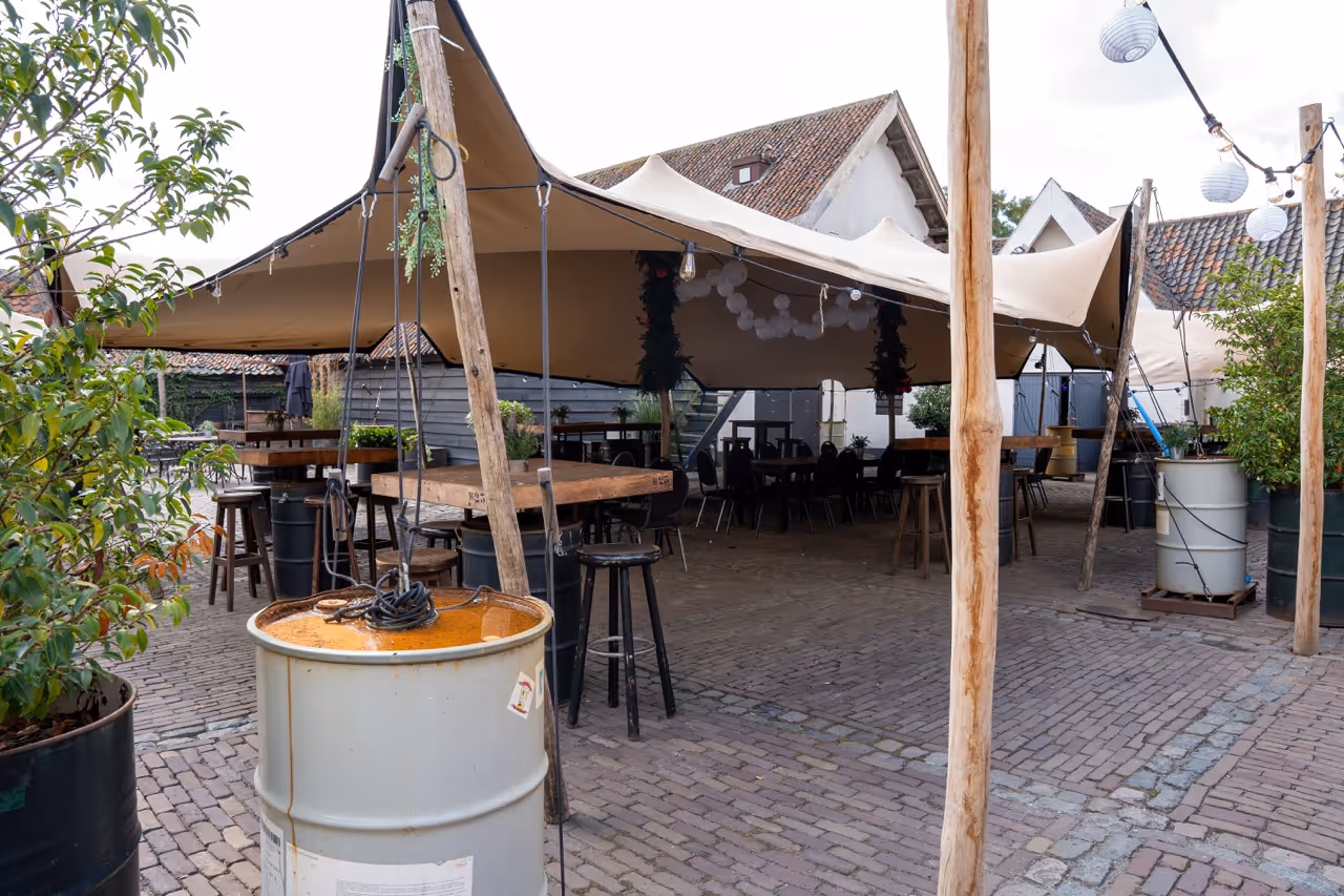 Outdoor seating area with wooden tables and stools under a large beige canopy tent on a cobblestone patio.