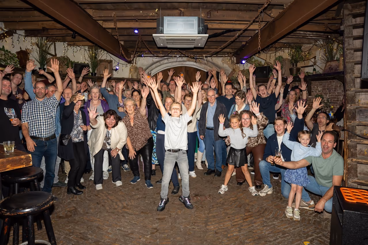 Large group of people of various ages raising their hands and smiling indoors under a wooden ceiling with string lights.