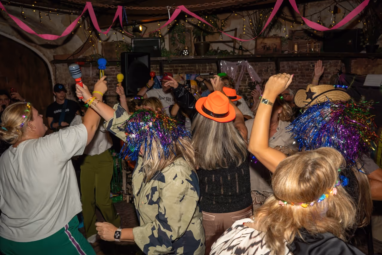 Group of people dancing and celebrating indoors with colorful hats, festive headbands, and raised hands.