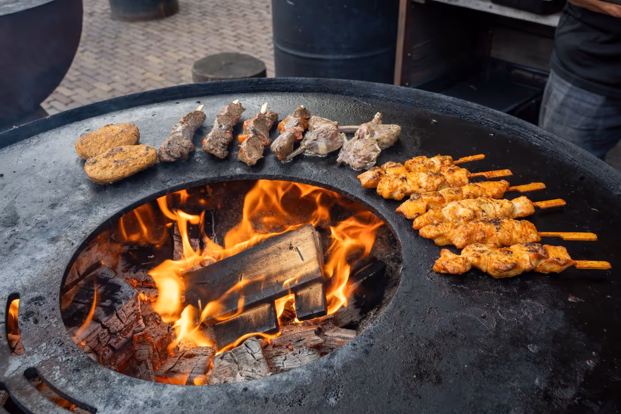 Skewers of meat and two patties grilling around a circular open-flame fire pit.