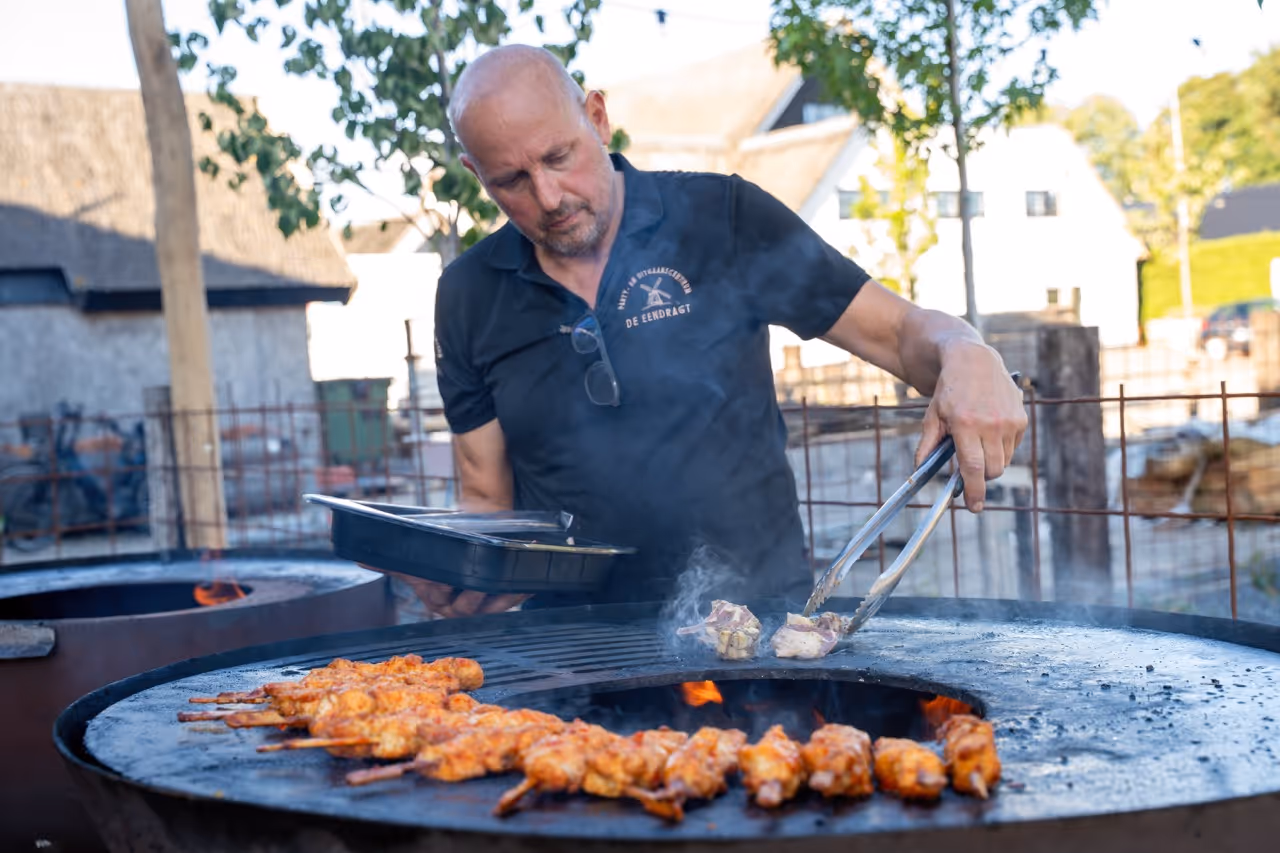 Man grilling skewers of marinated meat on a large round outdoor grill with smoke rising.