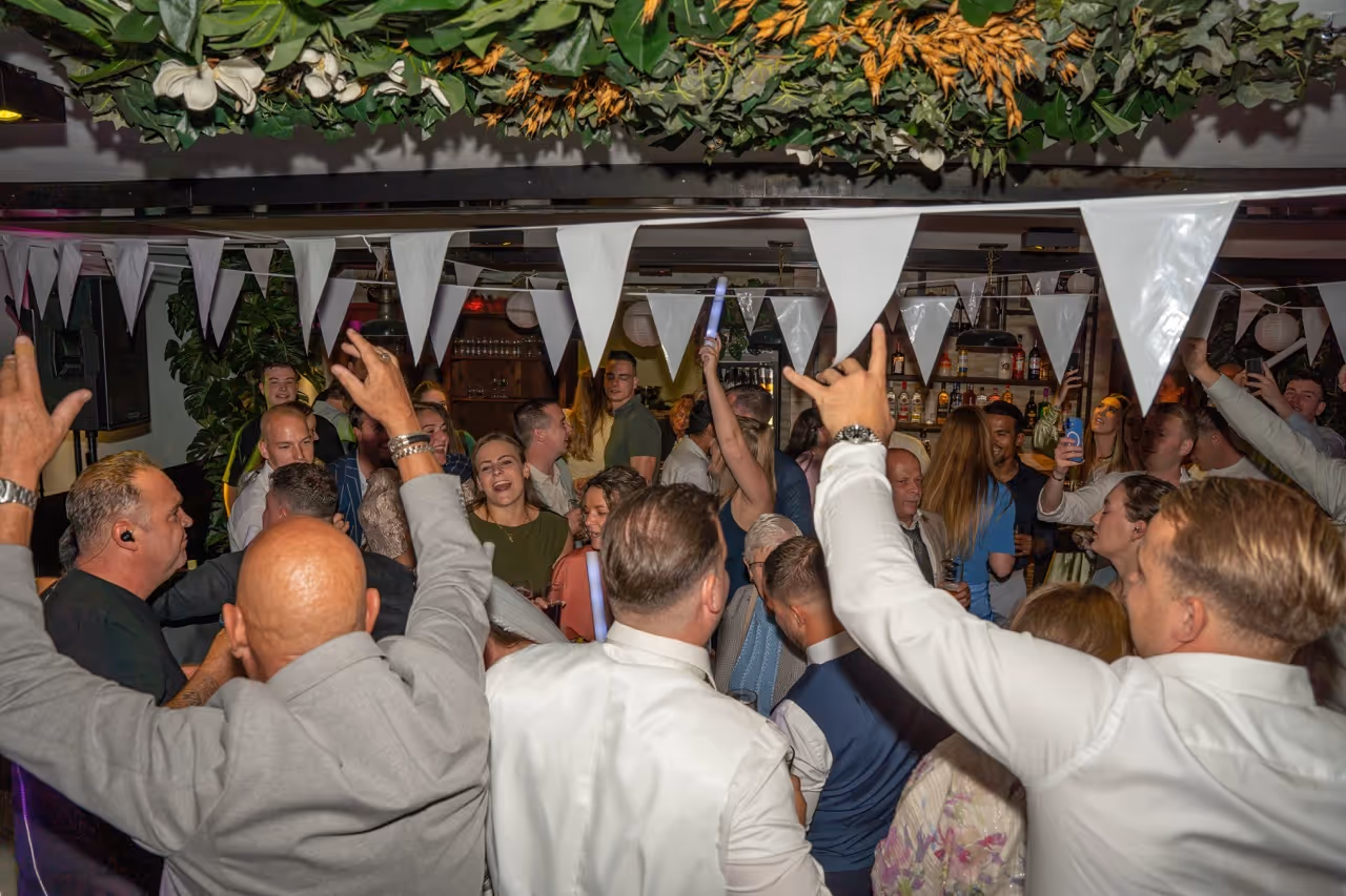 A crowded indoor party with people dancing and celebrating under white triangular bunting and green foliage decorations.