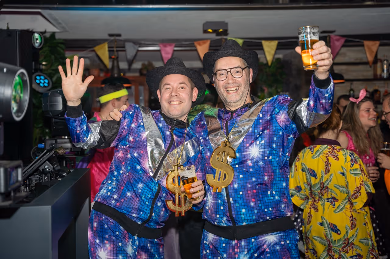 Two men in matching blue and silver disco outfits with large gold dollar sign necklaces, smiling and holding drinks at a lively party.