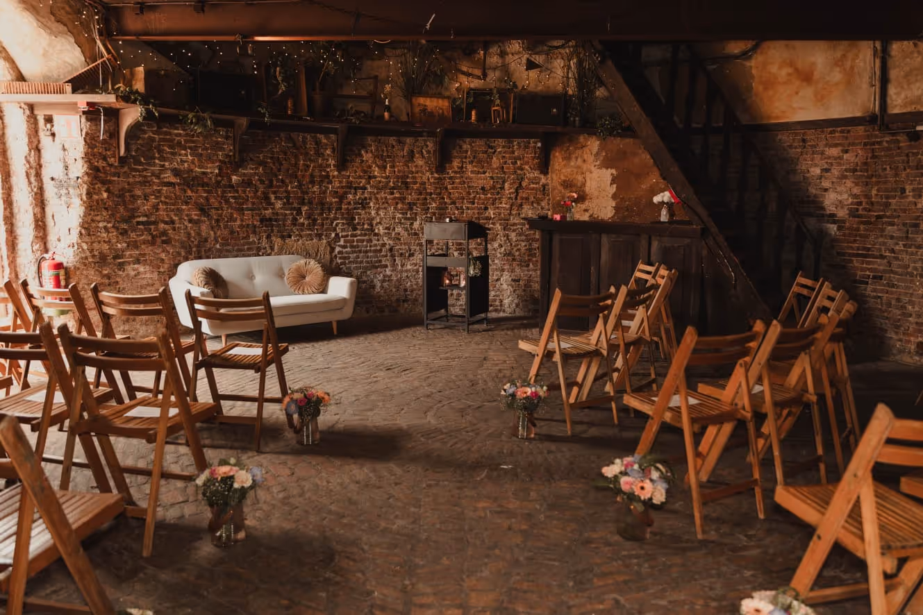 Cozy rustic room with wooden folding chairs arranged in rows, small floral bouquets on the floor, a white loveseat with round pillows, and exposed brick walls.