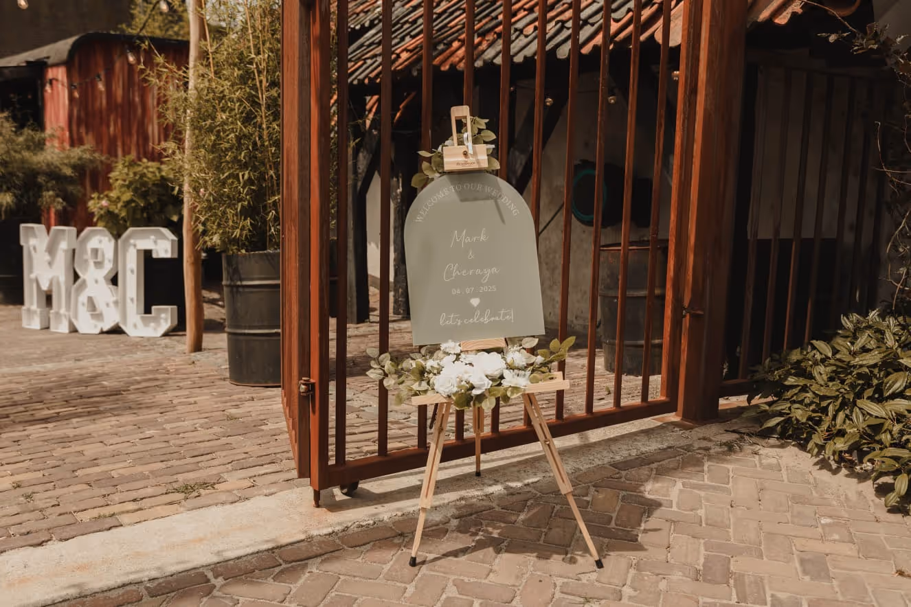 Wedding welcome sign on wooden easel decorated with white flowers, with a metal gate and large 'M&C' letters in the background.
