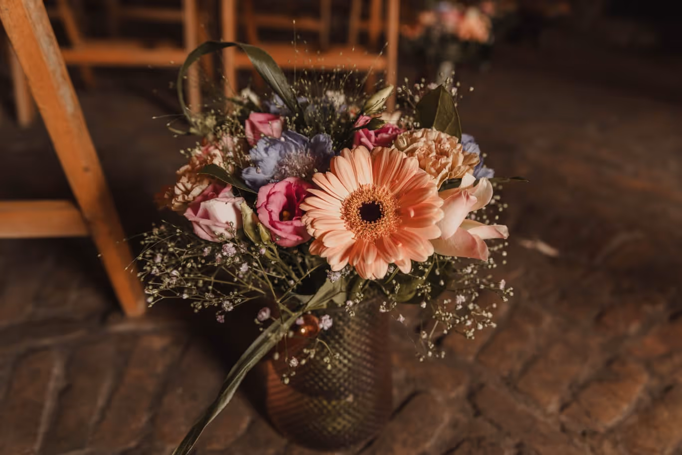 Close-up of a mixed flower bouquet in a textured vase on a stone floor with wooden chairs in the background.