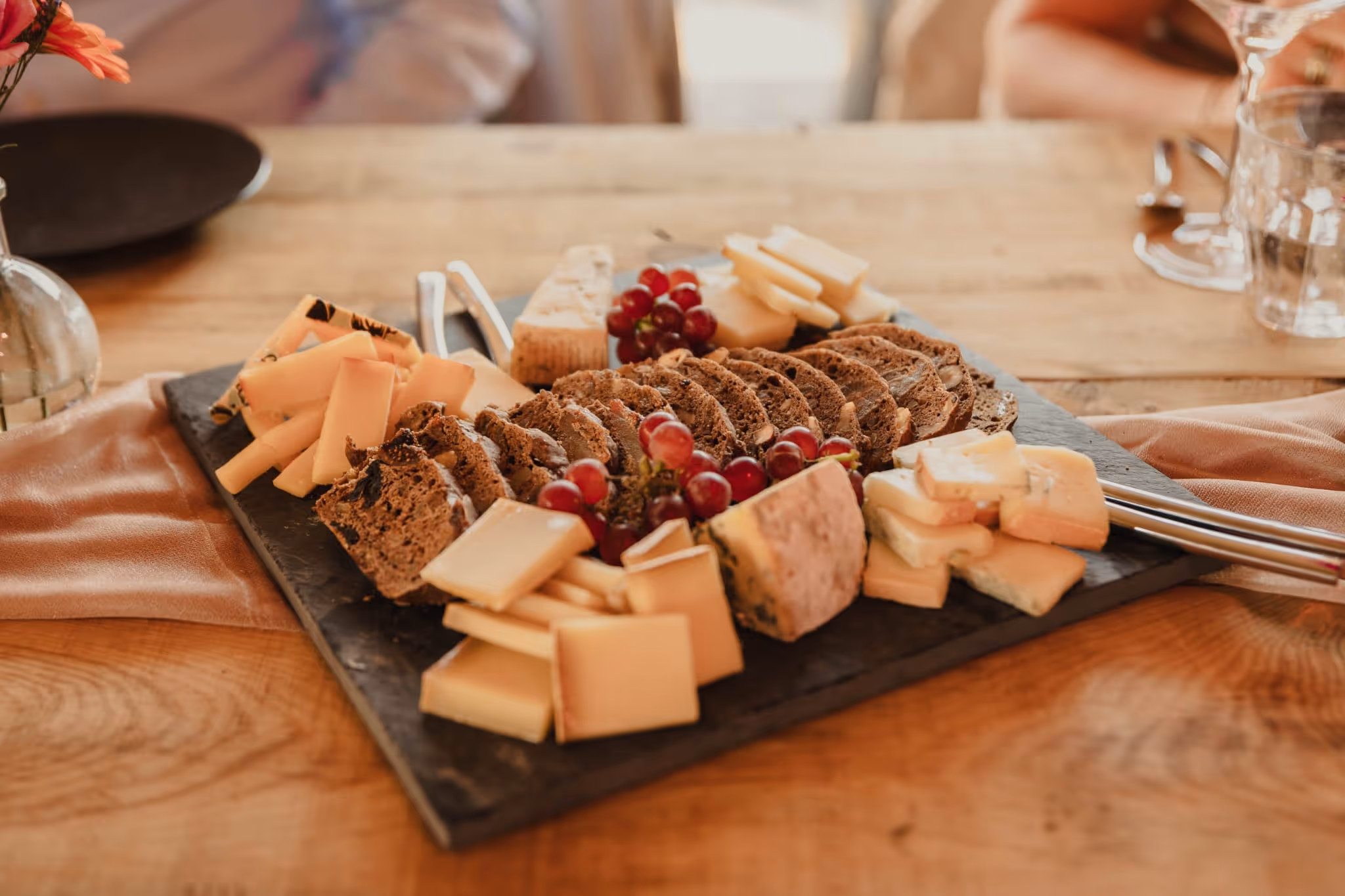 Slate platter with sliced bread, assorted cheeses, and red grapes on a wooden table.