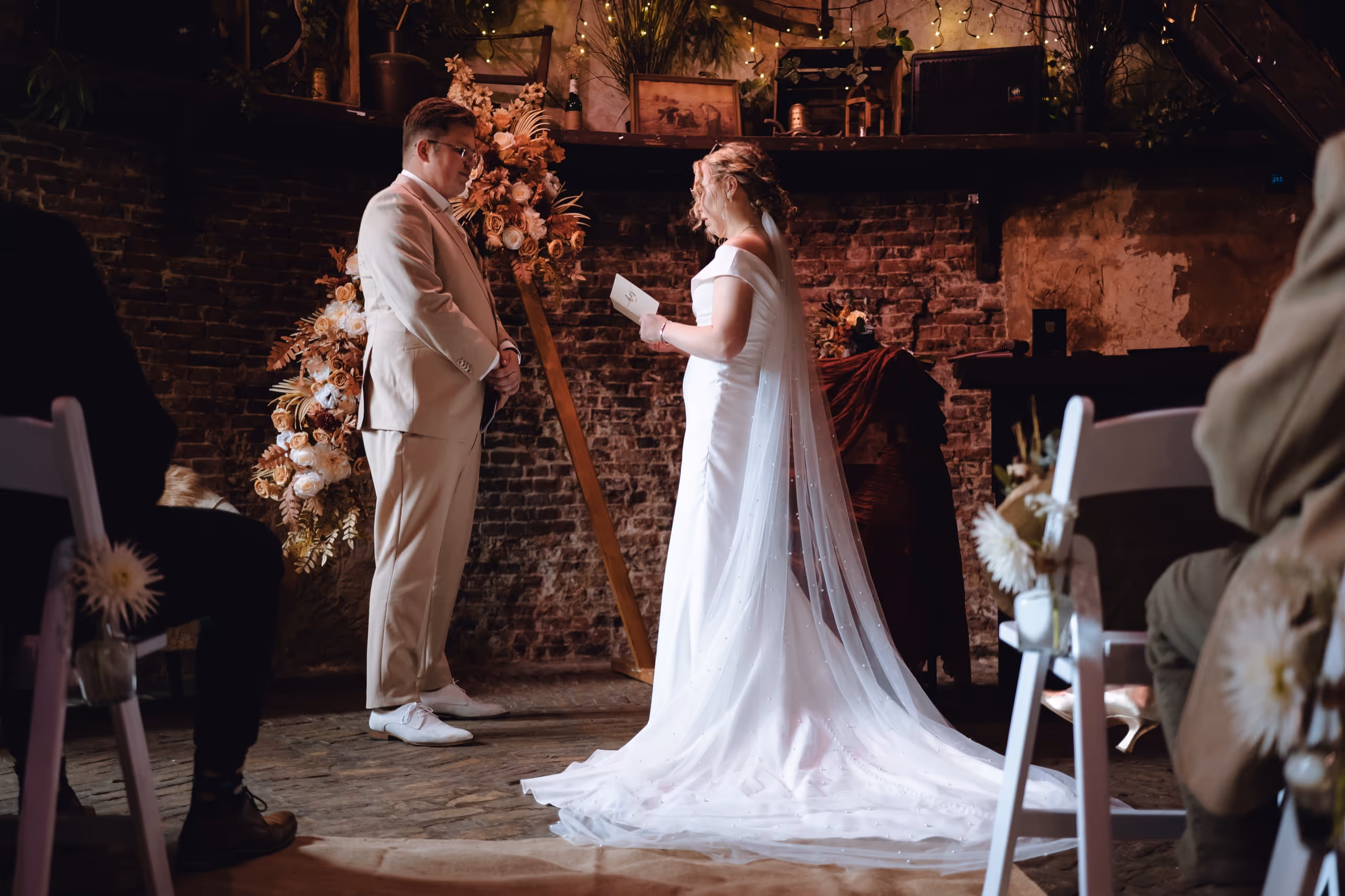 Bride in a white gown with a long veil reading vows to groom in a light beige suit during an intimate indoor wedding ceremony.