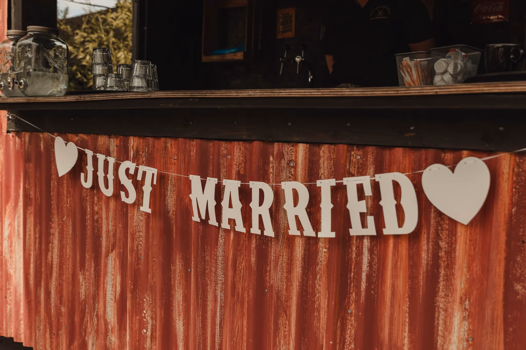 White 'JUST MARRIED' banner with hearts hanging on a rustic red corrugated metal wall.