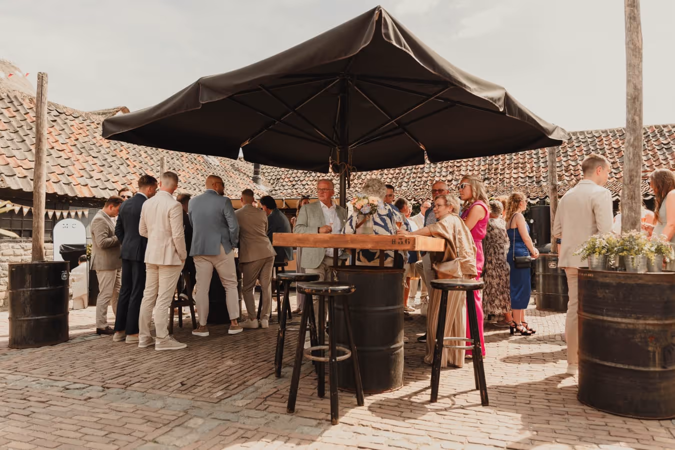 Group of people socializing outdoors under a large black umbrella with high tables and stools on a sunny day.