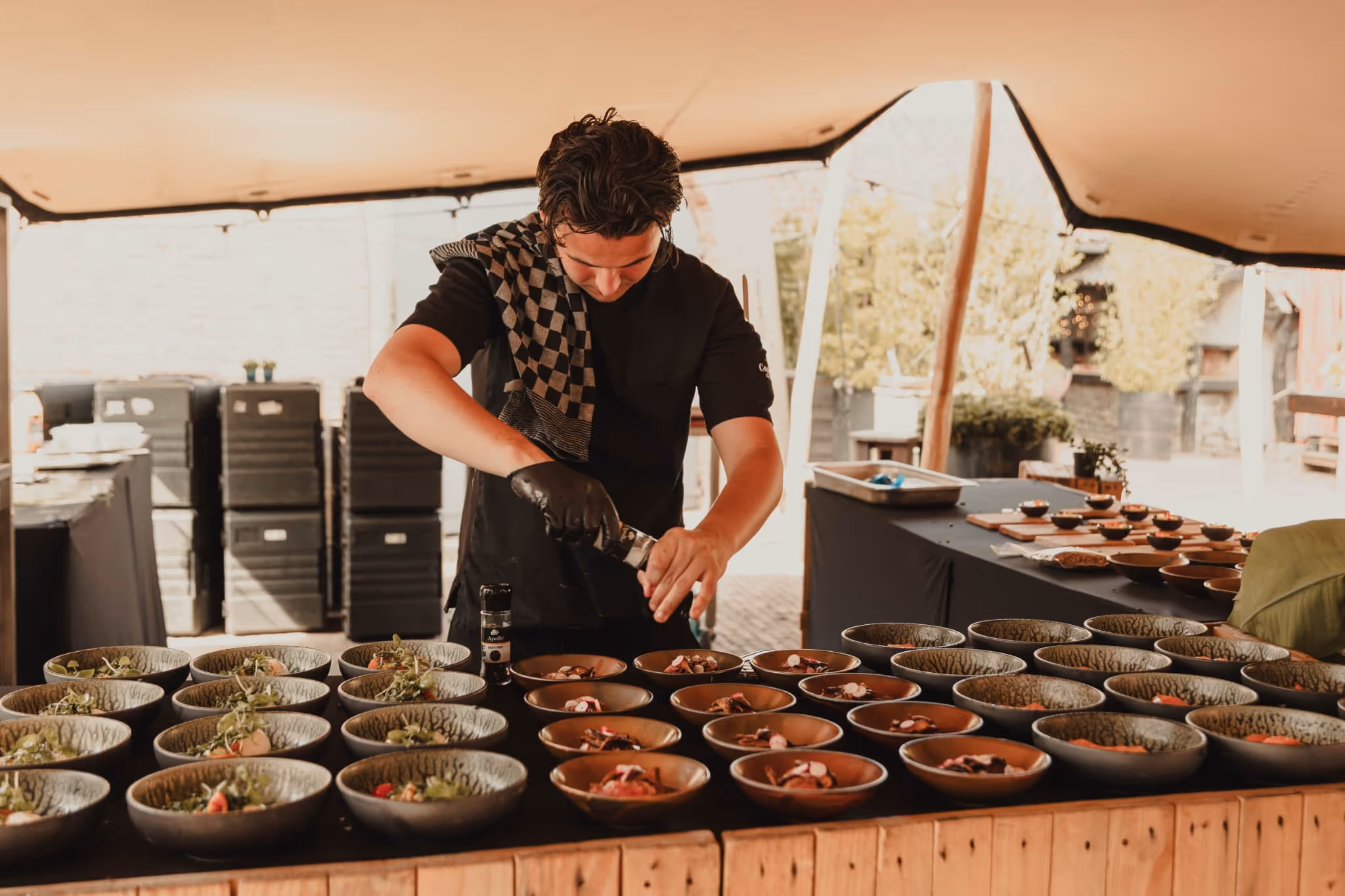 Chef wearing black shirt and gloves seasoning multiple bowls of food arranged on a wooden table under a tent.