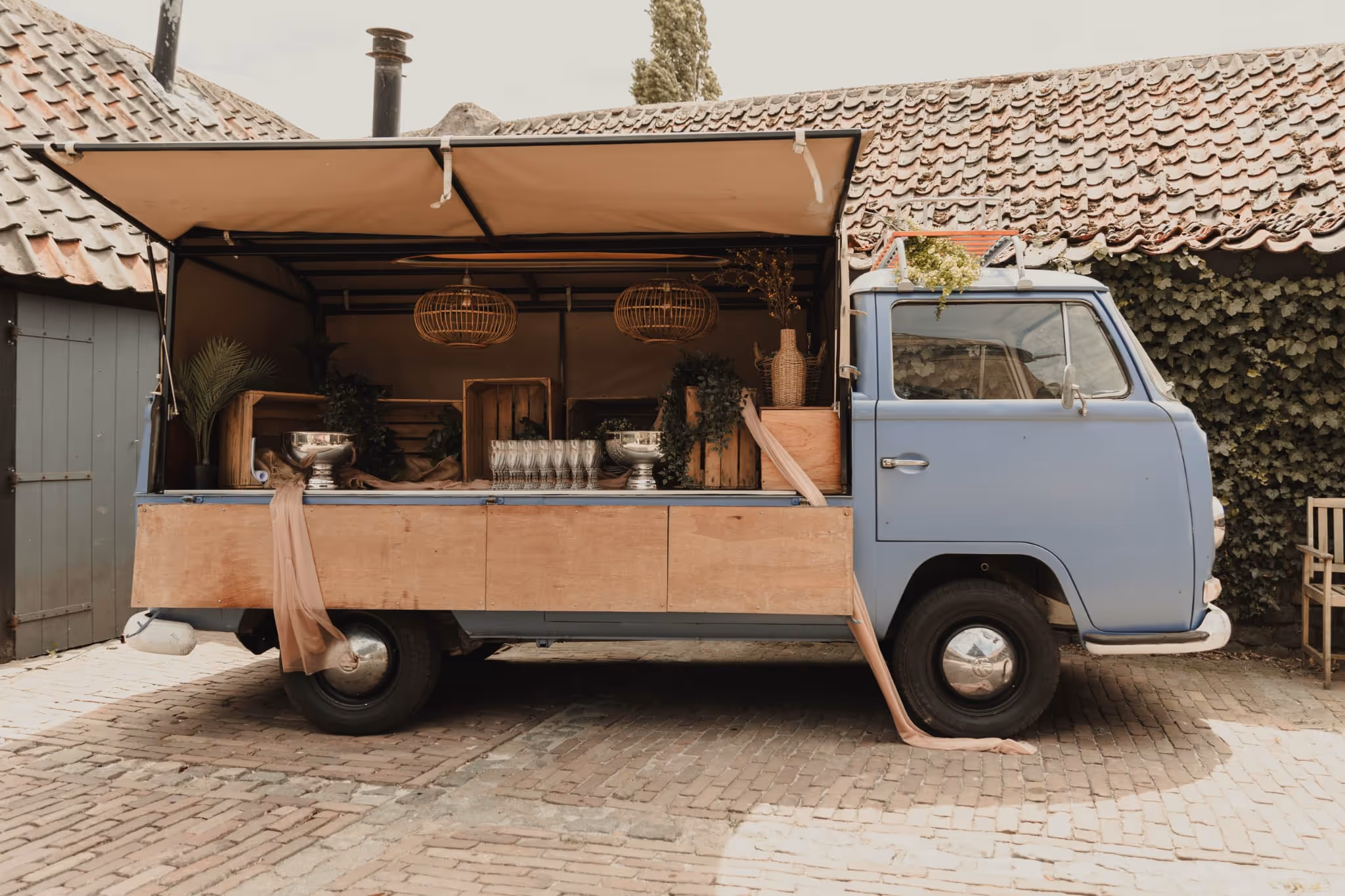 Light blue vintage van converted into a rustic mobile bar with wooden crates, glassware, greenery, and hanging wicker lamps.