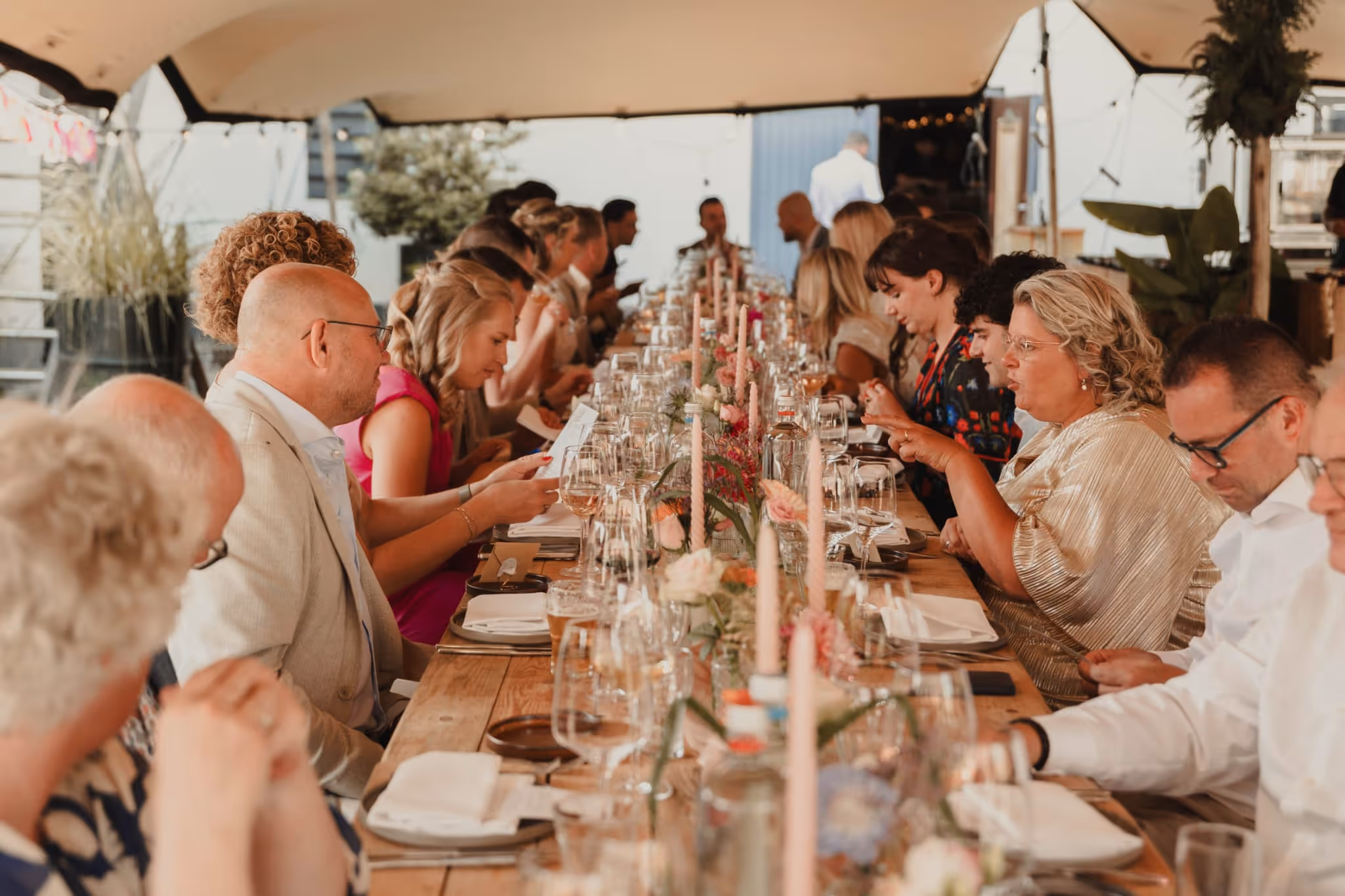 Long wooden table with guests seated on both sides under a tent, set with candles, flowers, and glassware for a formal event.