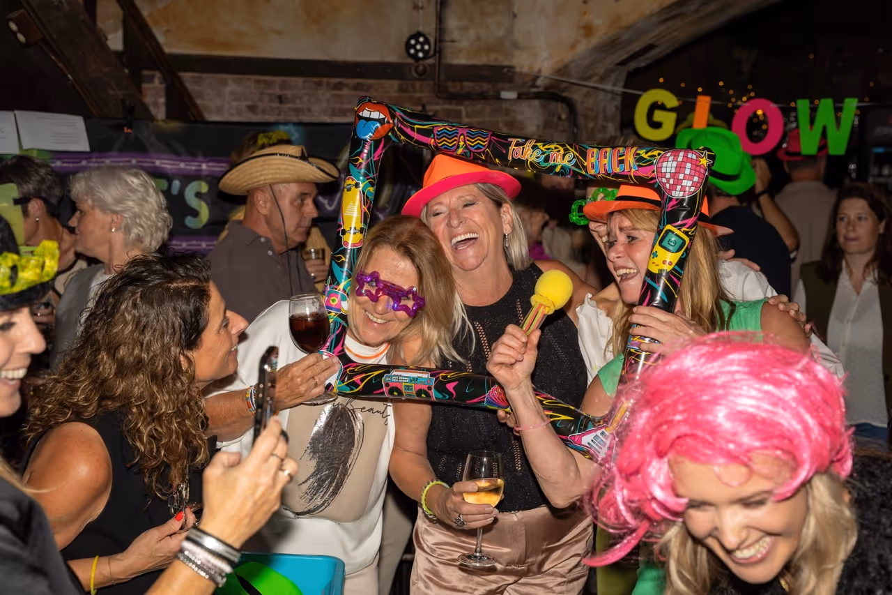 Groep mensen viert feest met kleurrijke accessoires zoals een roze pruik, hoeden, en een opblaasbare fotolijst met teksten.