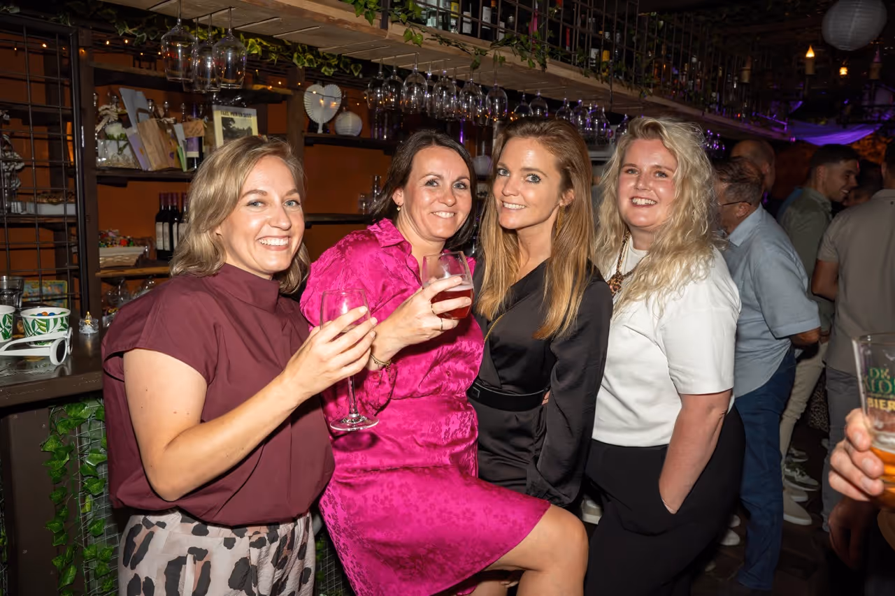 Vier vrouwen lachen en poseren met drank in de hand in een gezellige baromgeving.