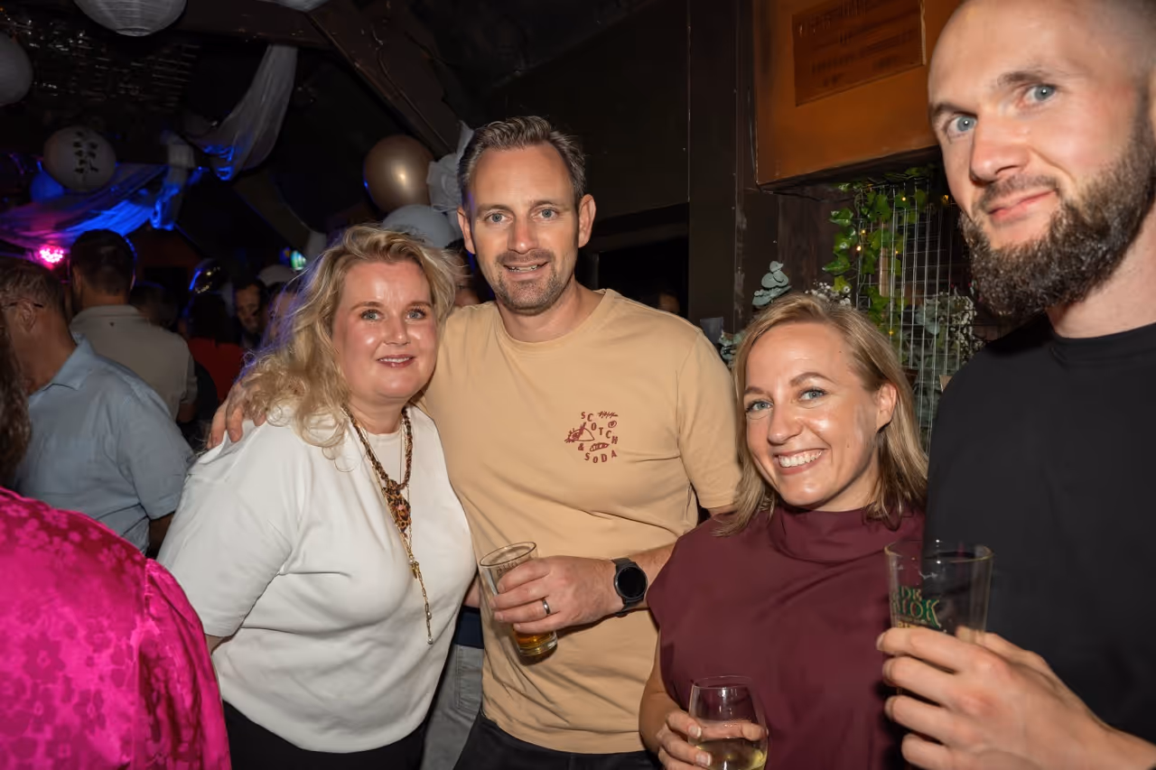 Vier mensen glimlachend en poseren samen met drankjes in een feestelijke omgeving.