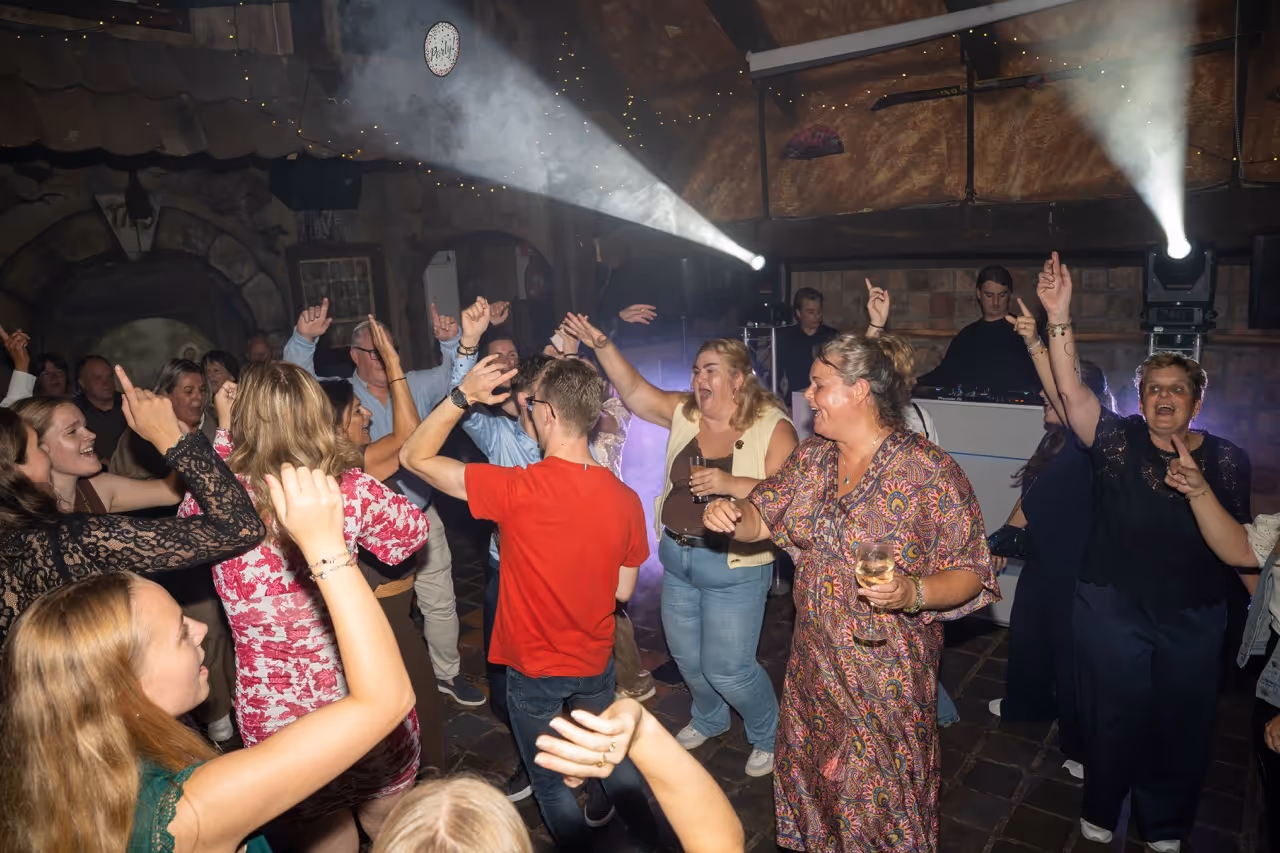 Mensen dansen en genieten op een feest met lichteffecten en een dj in een rustieke ruimte.