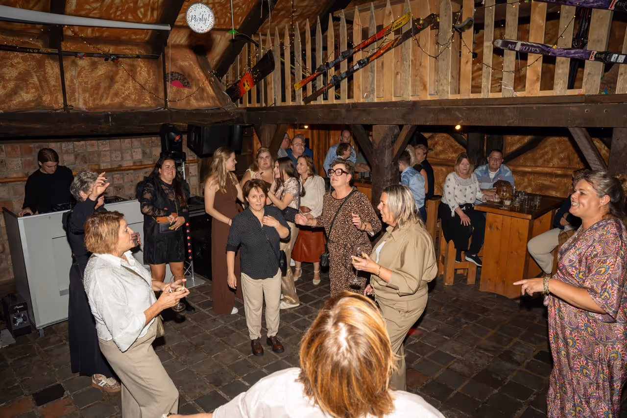 Groep mensen die in een rustieke ruimte met houten balken en ski-uitrusting aan de muur een feest vieren en dansen.