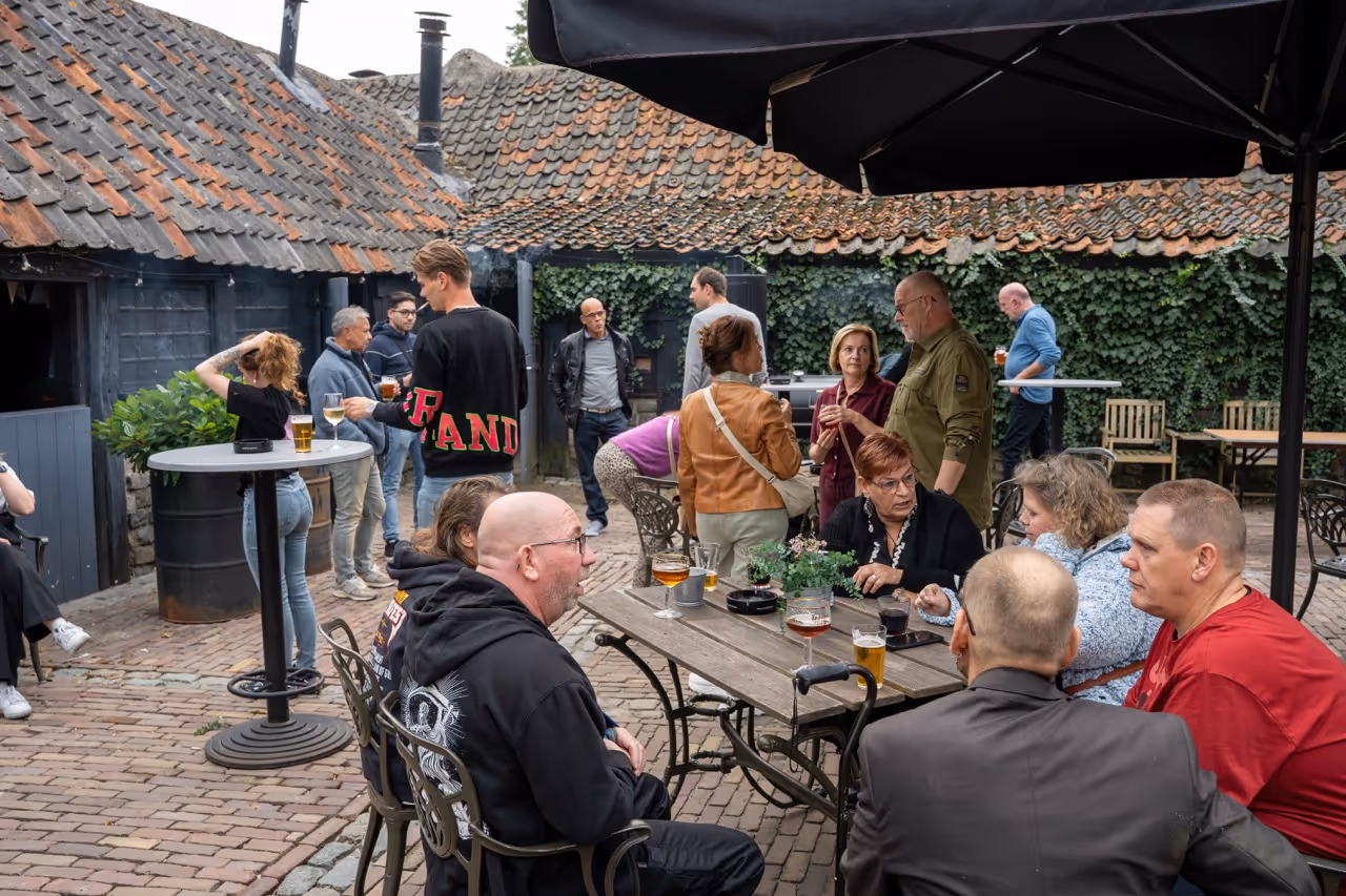 Groep mensen die buiten in een patio met stenen vloer rond houten tafels en hoge tafels sociale gesprekken voeren terwijl ze drankjes drinken.
