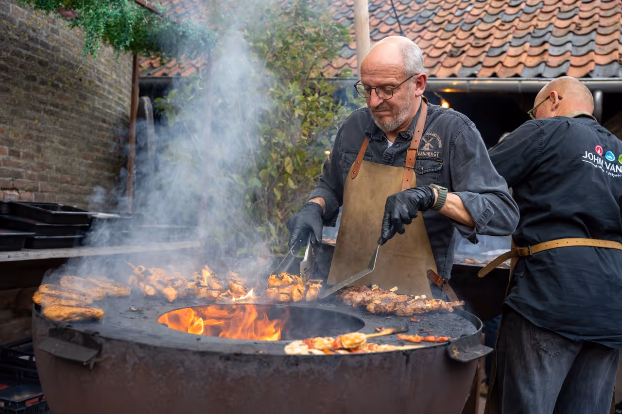 Twee mannen bereiden gegrilde spiesjes en kip op een grote open barbecue met vuur en rook.