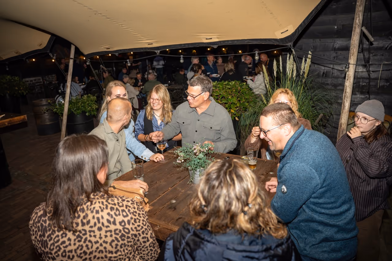 Groep mensen zit lachend rond een houten tafel onder een tent, genietend van een avond samen.