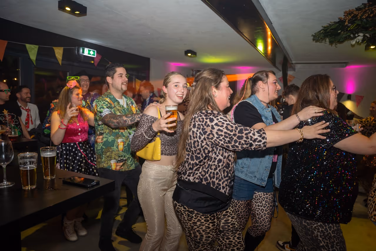 Groep mensen in feestelijke kleding en luide patronen dansen in een rij binnenshuis tijdens een feest.
