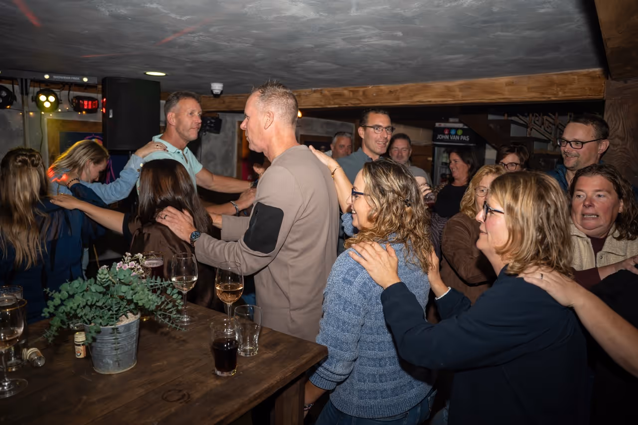 Groep mensen die in een kring staan met handen op elkaars schouders tijdens een sociale bijeenkomst binnenshuis.