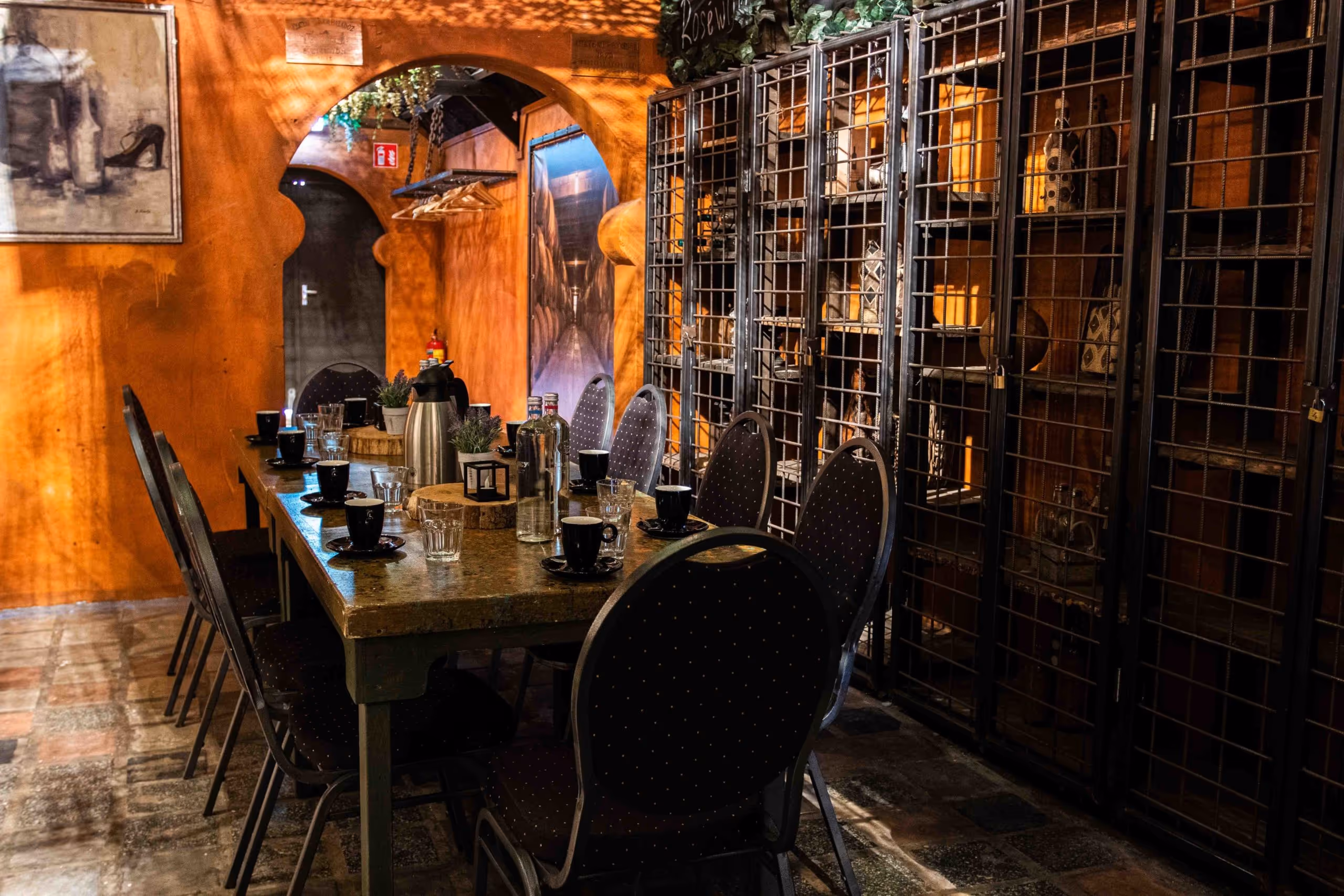 Dining room with long table set with black cups, glasses, water bottles, and thermos, surrounded by chairs and metal cage shelves filled with pottery.