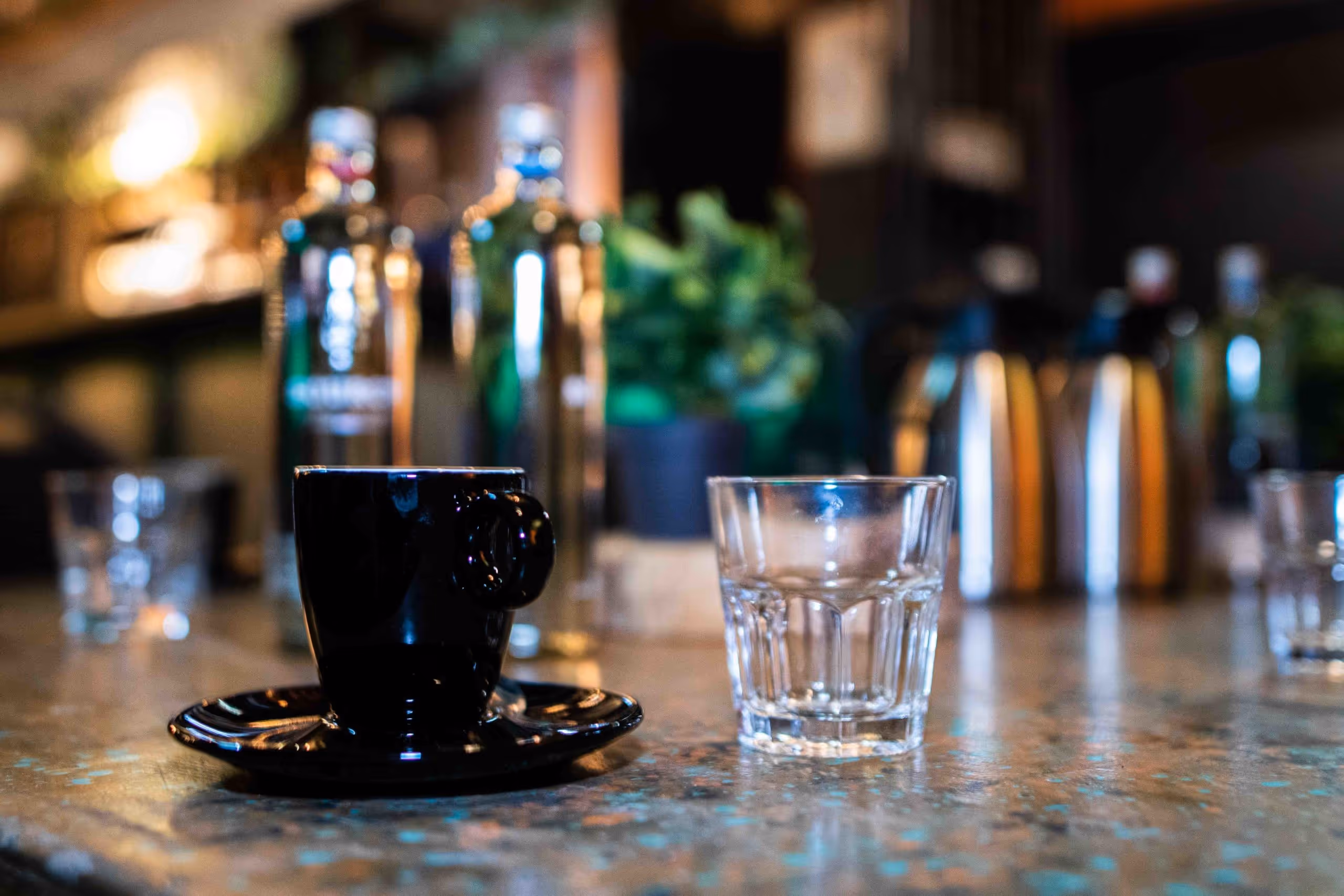 A black espresso cup and a clear glass placed side by side on a textured countertop with blurred bottles and a potted plant in the background.