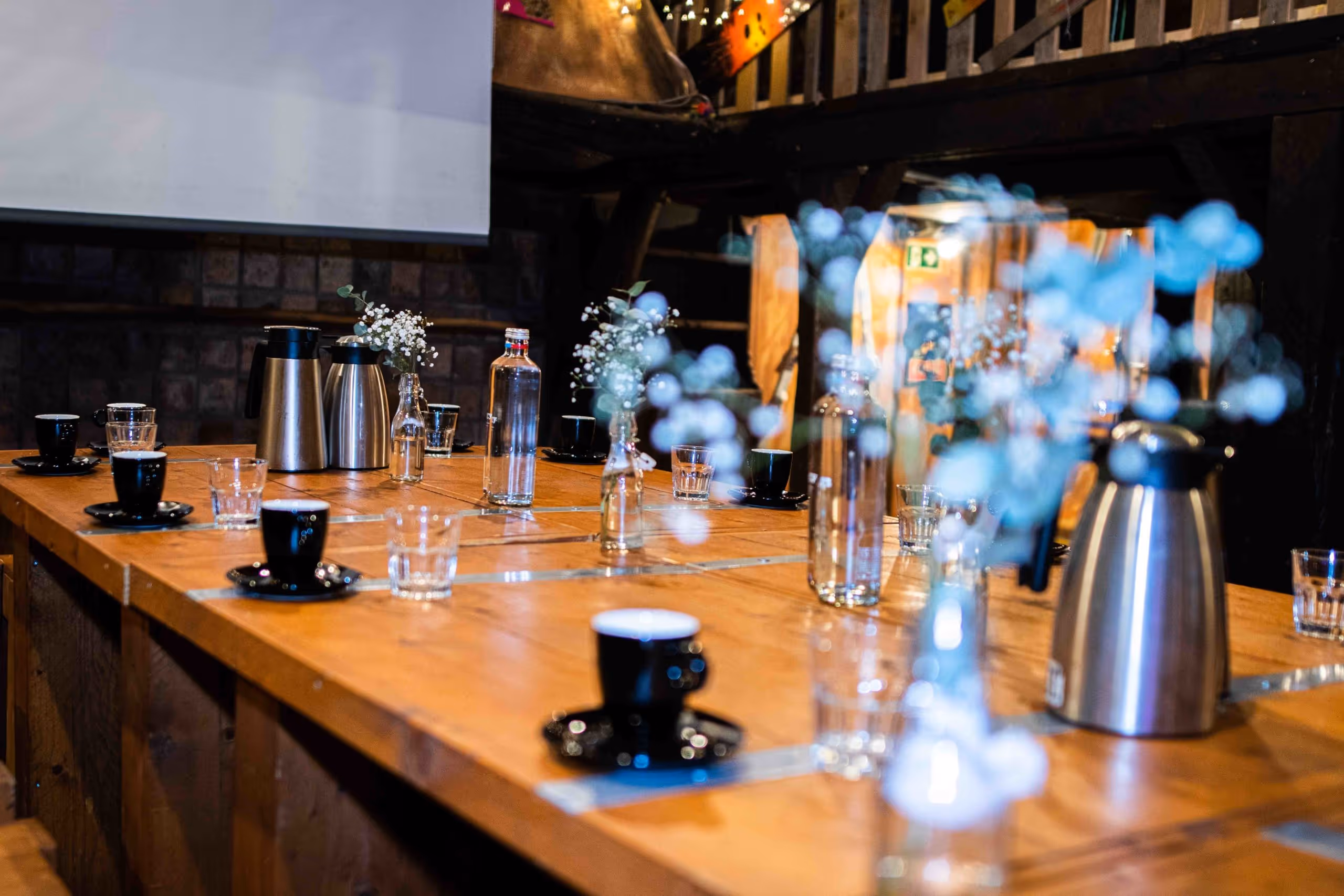 Wooden table set with black coffee cups, glass water bottles, tumblers, and small vases of white flowers in a cozy indoor setting.