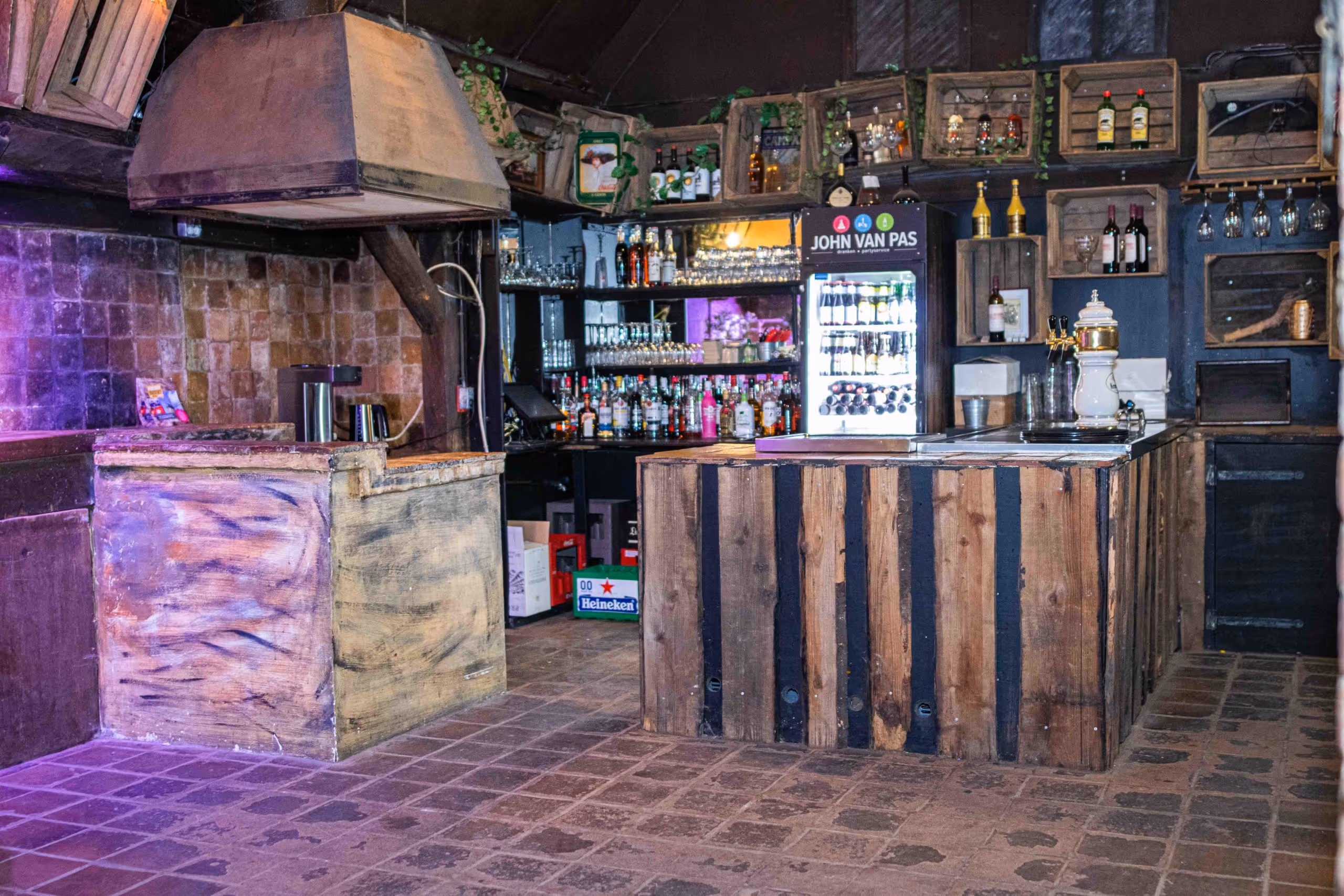 Rustic bar interior with wooden counters, shelves holding liquor bottles, glassware, and a refrigerator labeled John Van Pas.