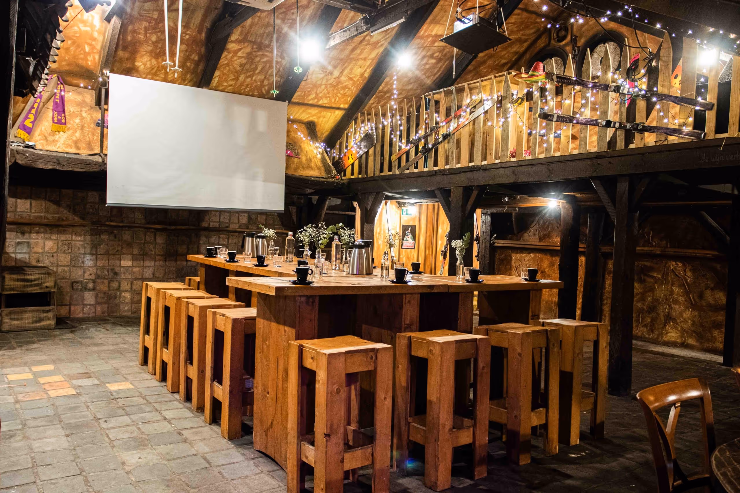 Cozy rustic room with wooden high tables and stools set with coffee cups and thermoses, decorated with string lights and skis on the balcony railing.