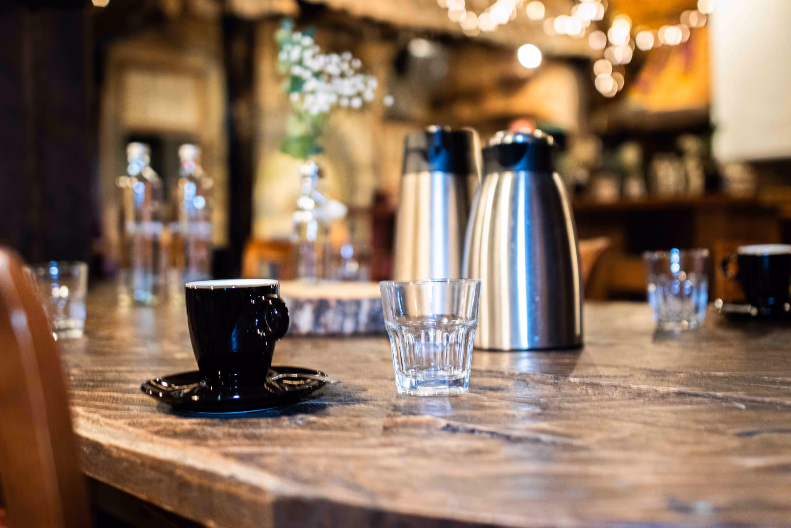 Close-up of a black coffee cup with saucer and an empty glass on a wooden table in a warmly lit café.