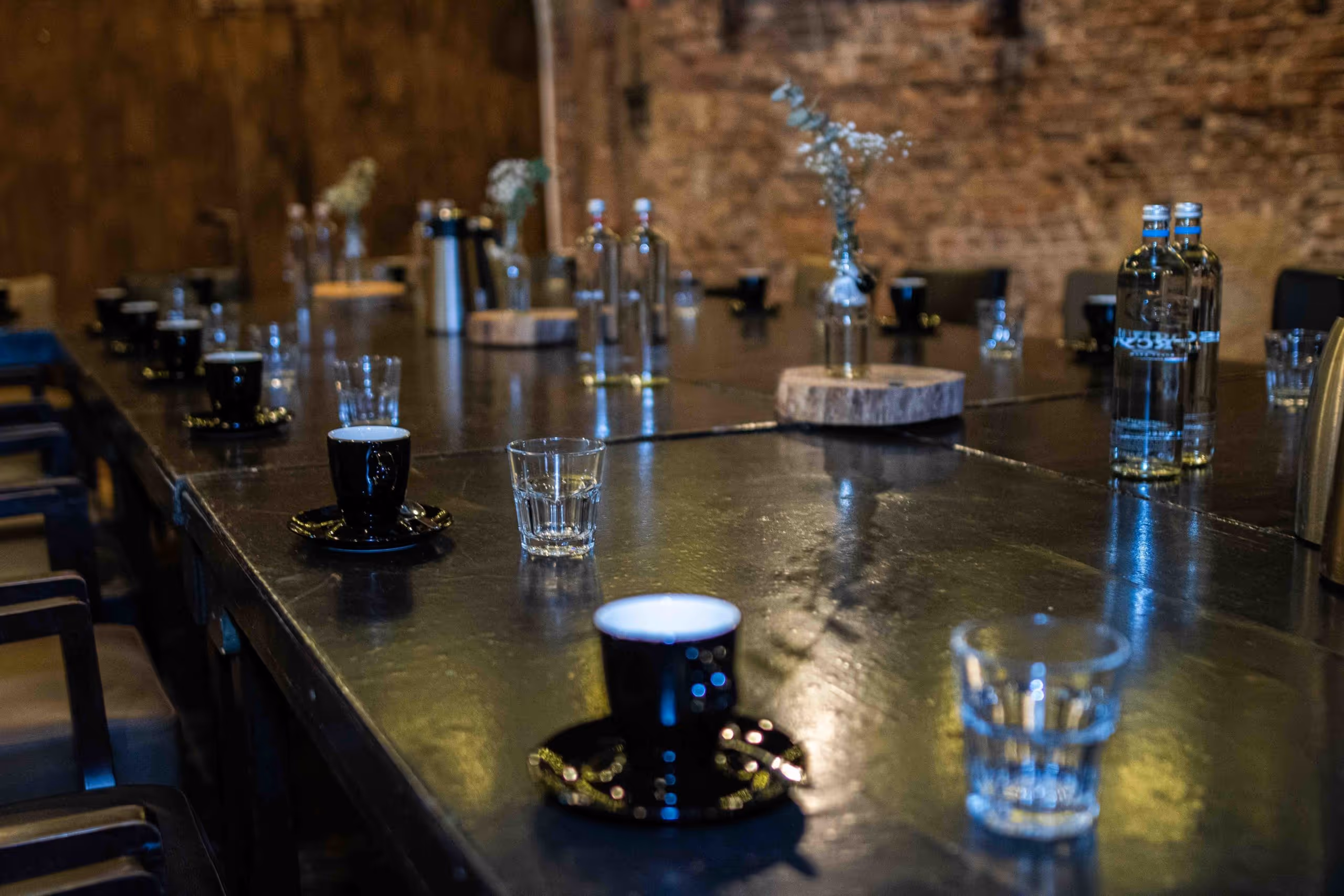 Long dark conference table set with black coffee cups, clear water glasses, bottles, and small vases with white flowers in an industrial-style room with exposed brick walls.