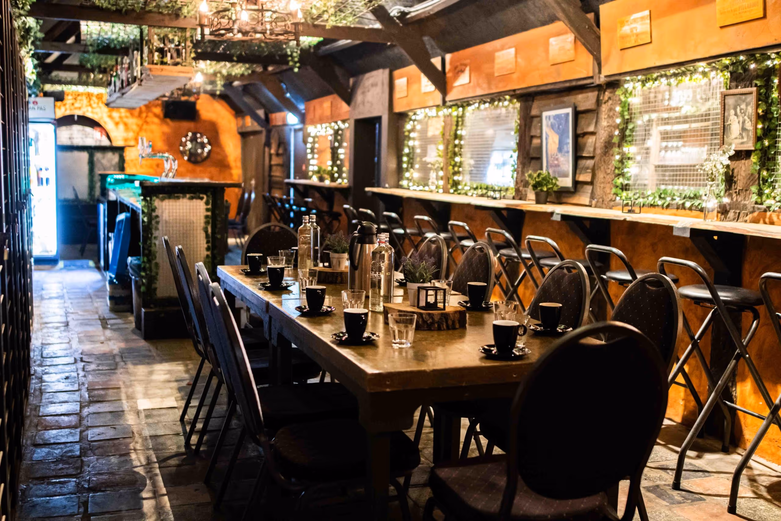 Cozy rustic cafe interior with a long wooden table set with black cups, glasses, bottles, and surrounded by chairs and bar stools along the wall decorated with lights and greenery.