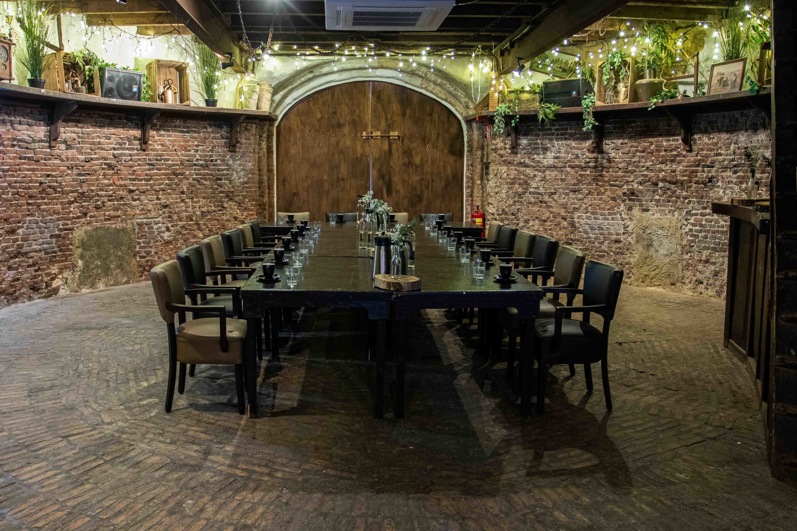 Long conference table with black chairs set in a rustic room with exposed brick walls, wooden ceiling beams, and string lights.