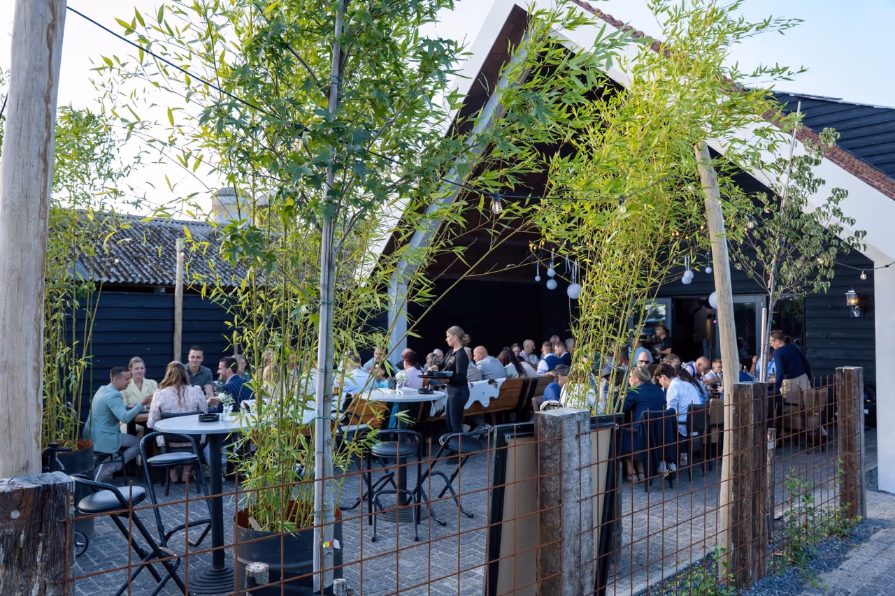 Outdoor restaurant patio with people dining under a peaked roof surrounded by greenery and bamboo plants.