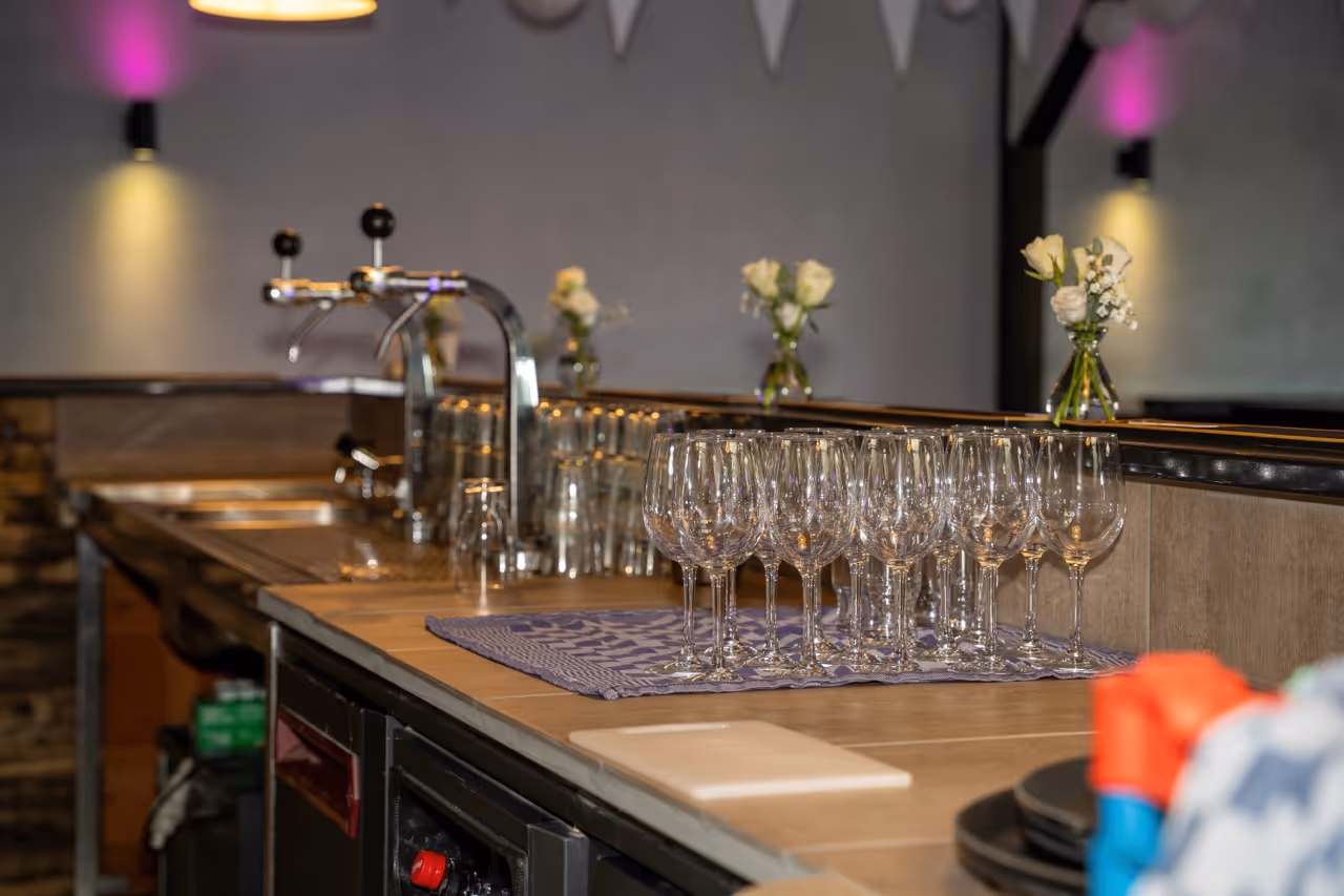 A bar counter with multiple empty wine glasses arranged on a cloth, beer taps in the background, and small vases of white flowers.