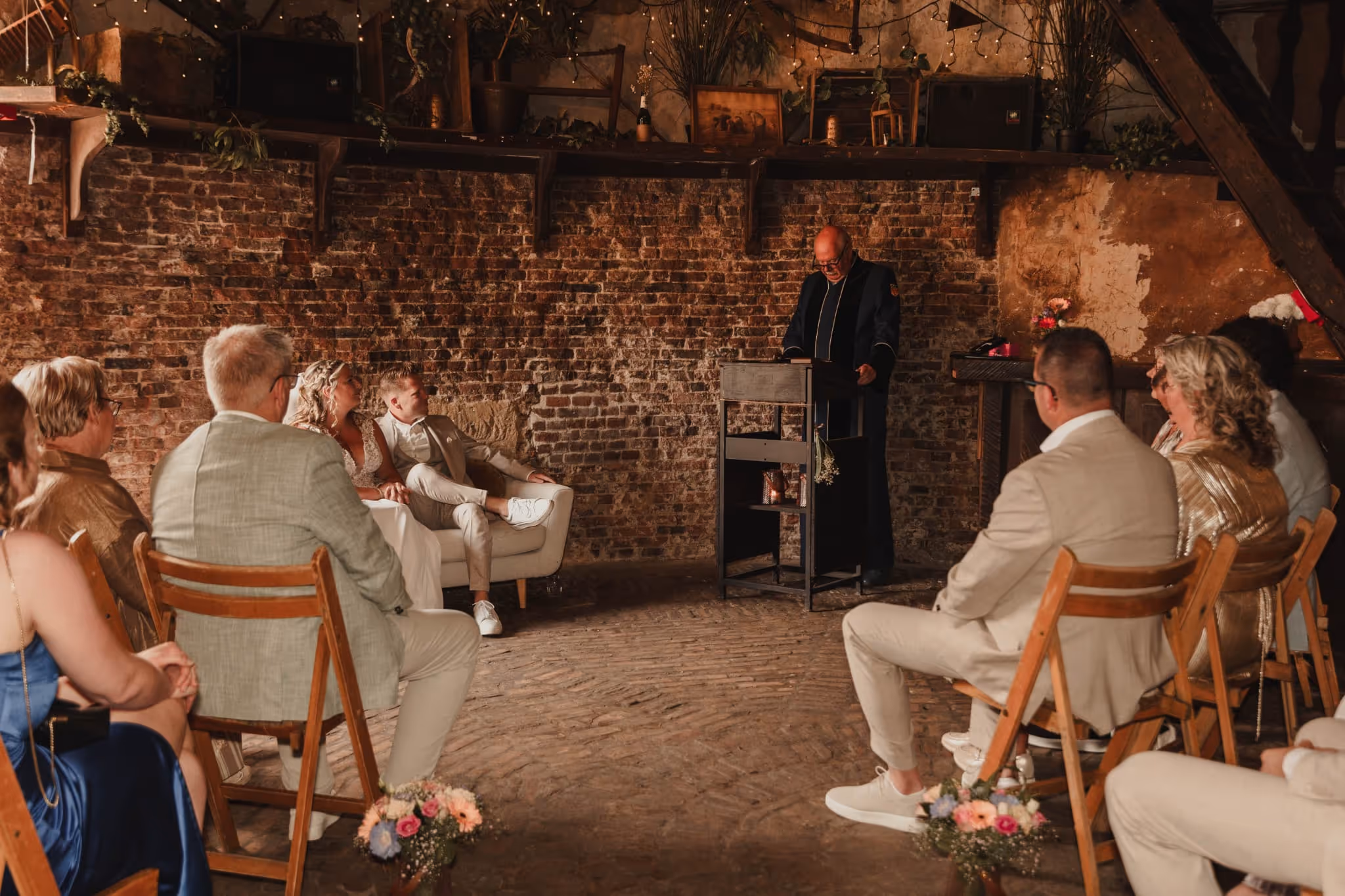 Een ceremonie in een rustieke ruimte met bakstenen muren waar een man een toespraak houdt voor een groep zittende gasten, inclusief een bruidspaar op een bank.