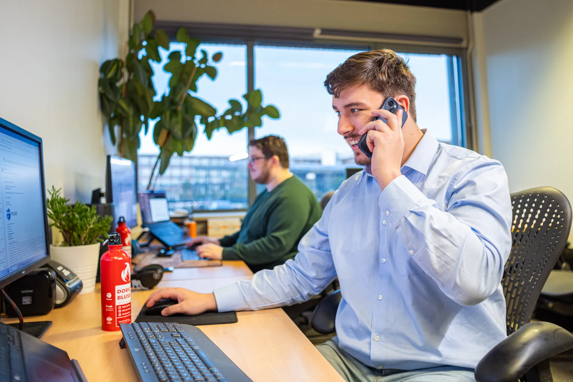Twee mannen zitten op kantoor; één belt terwijl hij naar zijn computerscherm kijkt, de ander werkt op een laptop bij het raam.