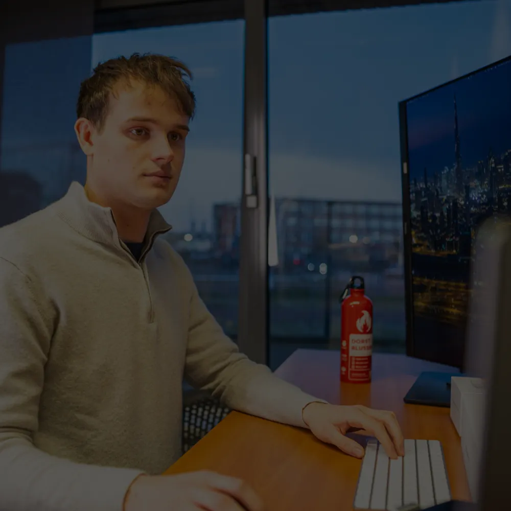 Jonge man zittend aan bureau met computer en waterfles, met avondstad op scherm achter hem.