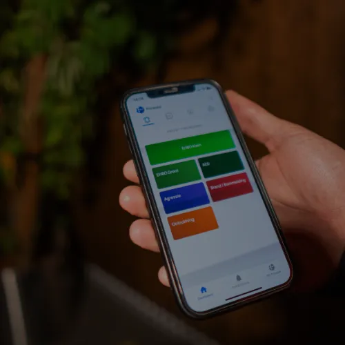 Hand houdt een smartphone met een app-scherm met gekleurde knoppen voor meldingen zoals EHBO-oefen, BRAND, agressie en ontruiming.